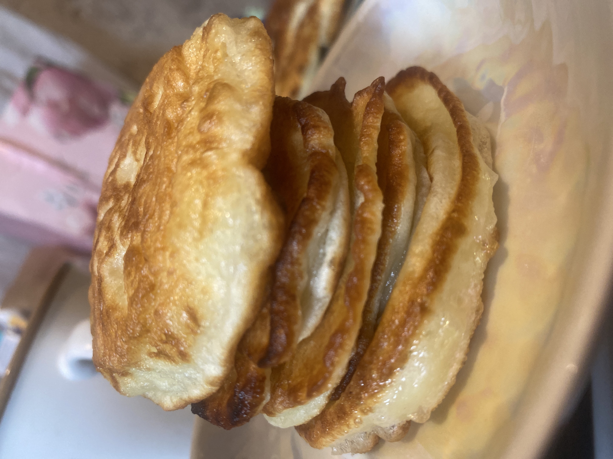 The image is a close-up, shallow depth of field photograph of a stack of golden-brown, fluffy pancakes on a white, iridescent plate. The pancakes are unevenly stacked, with visible air pockets and slightly browned edges. In the background, out of focus, a white mug or cup is visible to the left, and a pink object with floral patterns is to the right. The overall lighting suggests an indoor setting, possibly a kitchen or dining area. The image does not contain any information about the city of Kolomna, Russia.