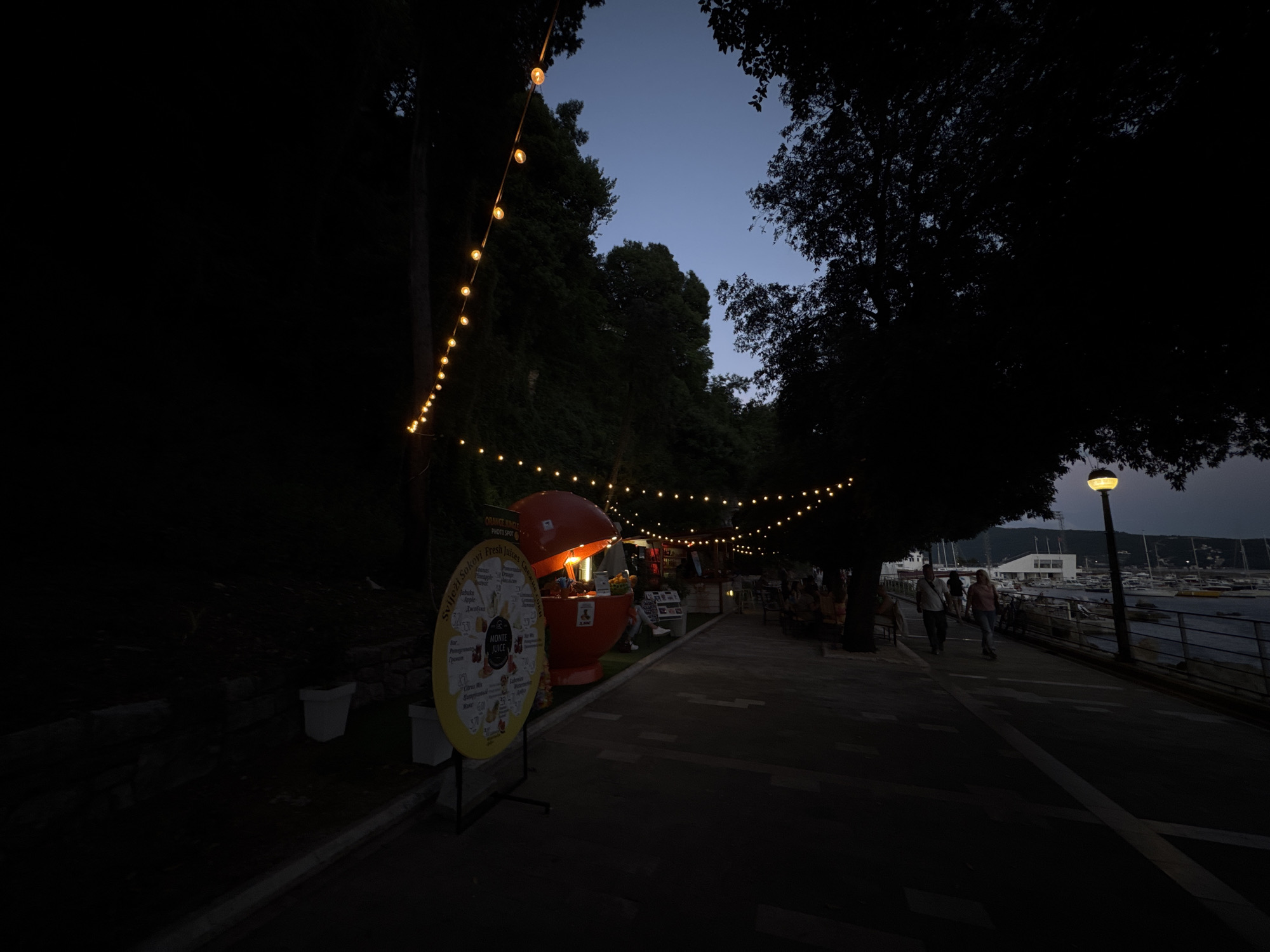 The image shows a dimly lit pedestrian walkway along the waterfront in Herceg Novi, Montenegro at dusk. String lights are strung above an orange-shaped juice stand, illuminated and open for business. A yellow sign with the words "MONTE JUICE" and "Sviježi Sokovi Fresh Juices Свежи" is placed in front of the stand. People are walking along the path toward the water, where boats are docked near a white building in the distance. A streetlight is visible on the right side of the walkway. The scene is framed by trees.