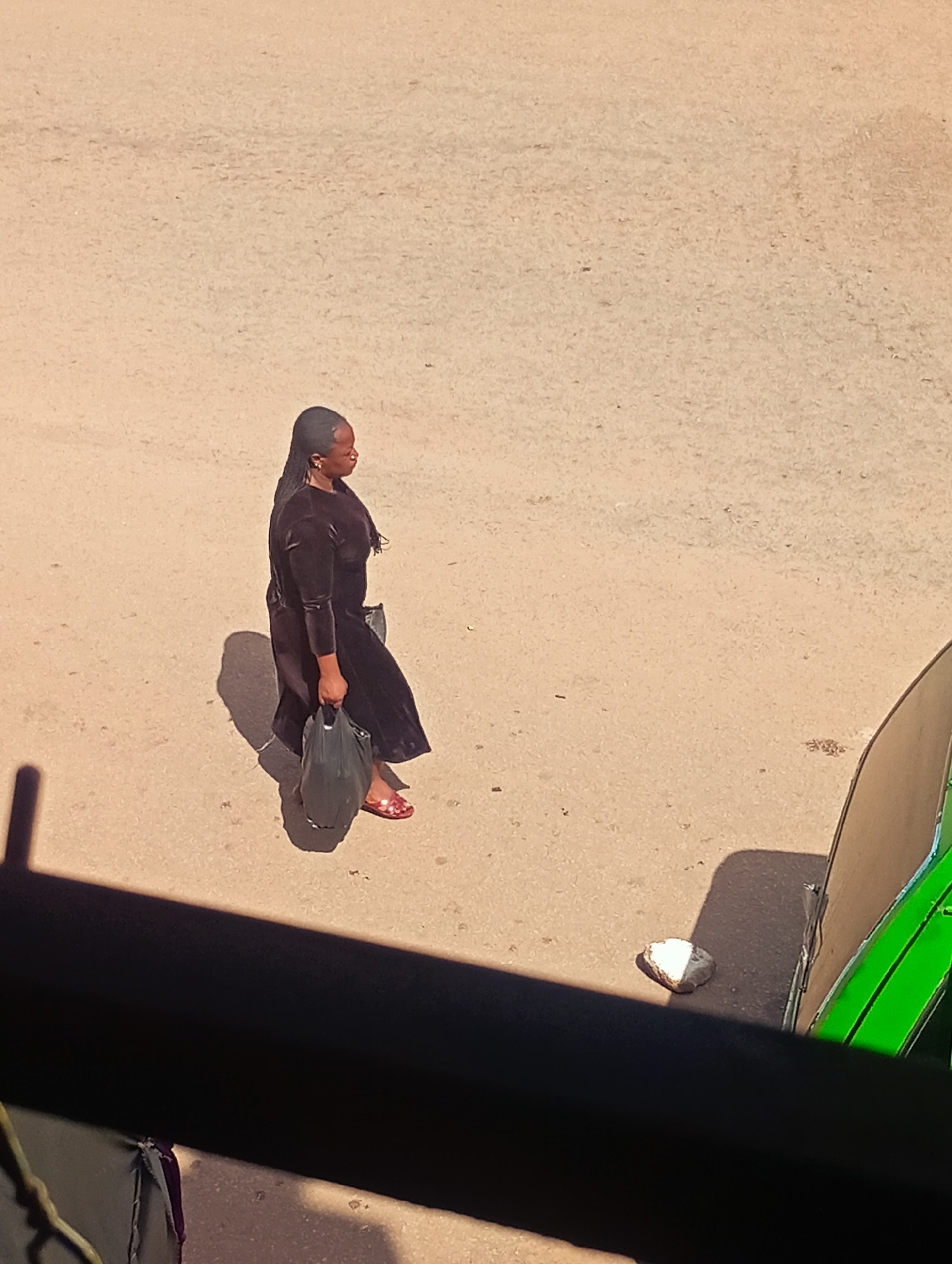 In Bukuru, Nigeria, a Black woman walks across a dusty, sun-baked surface, possibly a road or open area. She wears a long, dark, velvet dress and carries a large black plastic bag in her left hand. Her hair is dark and styled in braids that fall over her left shoulder. On her feet, she wears bright red flip-flops. A shadow stretches to her left, indicating the direction of the sun. In the lower right portion of the image, a green vehicle is partially visible, along with a small, light-colored rock. The overall environment appears dry and sparsely populated.