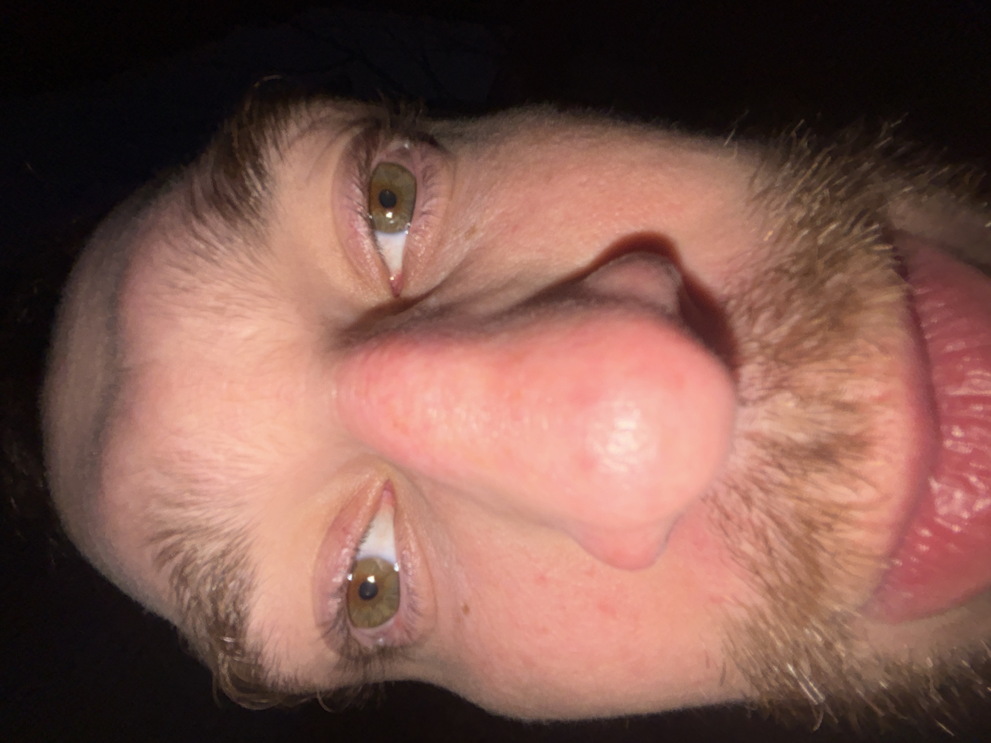 Close-up, rotated view of a person's face. The individual has light skin, hazel eyes, a large nose, and a reddish-pink tongue protruding slightly from their mouth. The person has a light brown beard and short brown hair. The background is dark and indistinct. There is no visible indication of the location being Krasnoyarsk, Russia.