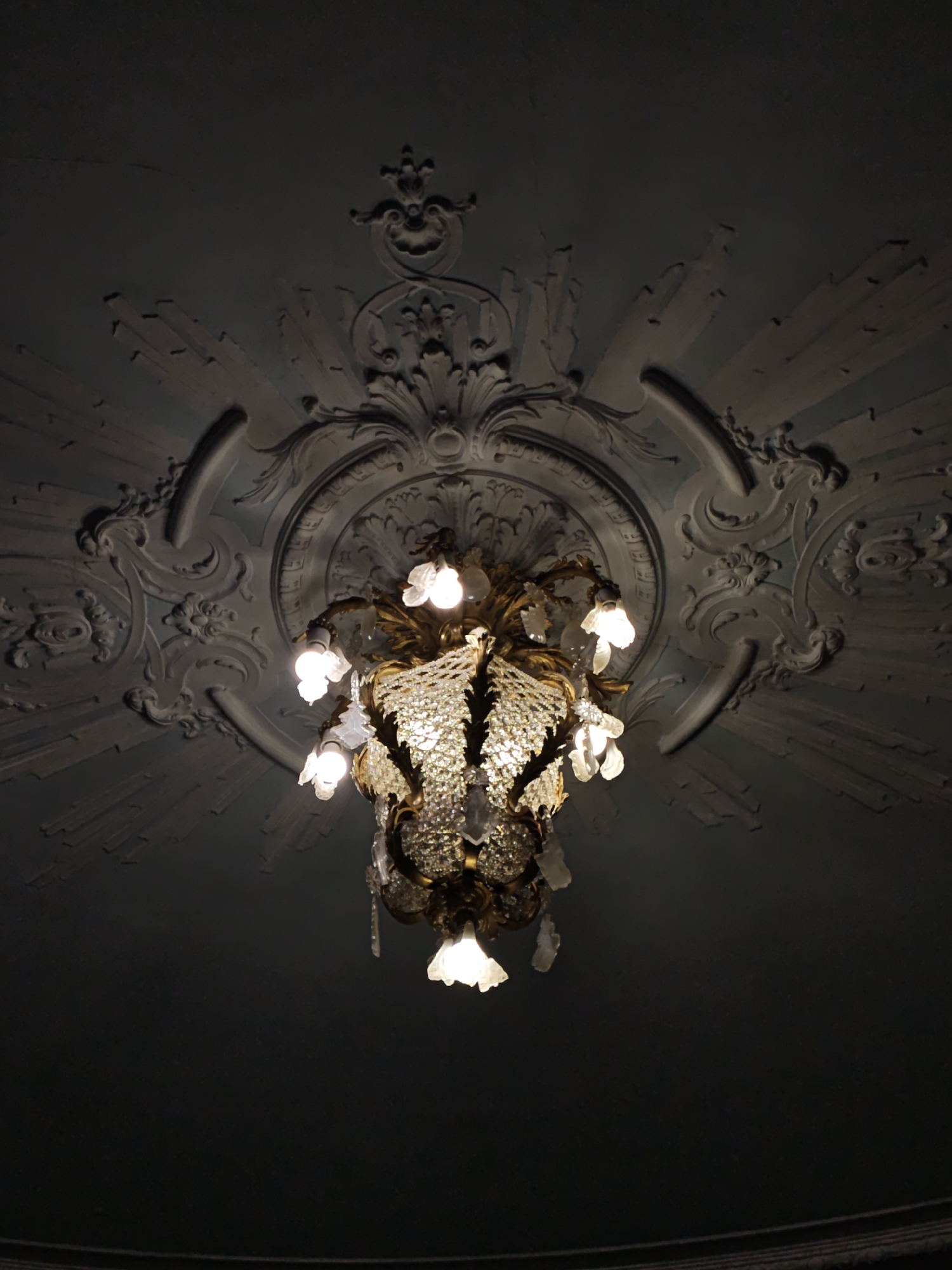 The image shows a chandelier hanging from a highly ornate ceiling in what appears to be an interior space. The ceiling features intricate plasterwork with a sunburst pattern radiating outwards from a central medallion, which is further adorned with elaborate scrolls and floral motifs. The chandelier itself is made of gold-toned metal and is heavily decorated with crystal elements that catch and reflect the light. Several bulbs are illuminated, casting a warm glow. The overall aesthetic suggests a classical or baroque style. The image does not contain any people or provide specific details about the location in Lviv, Ukraine.