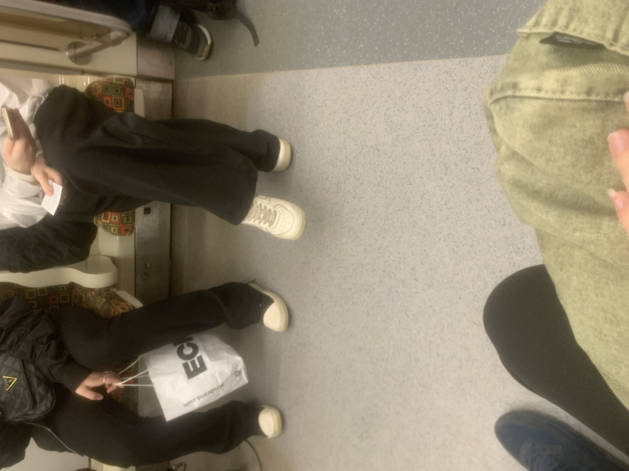 An image, rotated 90 degrees clockwise, captures a view inside what appears to be a public transport vehicle, likely a metro or train, from an overhead perspective. The floor is a light grey speckled material.
Several individuals are visible, mostly from the waist down. On the left side, three people are seated on upholstered seats featuring a distinctive geometric pattern of red, orange, green, and black squares and dots.
The uppermost seated person is wearing light-colored trousers and dark shoes with light soles. Below them, a person is wearing black wide-leg pants and off-white sneakers (resembling Nike Air Force 1s). This individual is holding a light-colored phone in one hand and a small white piece of paper in the other, with a white long-sleeved top visible. The lowest seated person on the left is also wearing black pants and off-white sneakers. They are holding a white shopping bag with visible text and have a black quilted bag, possibly a purse with a gold "G" logo, on their lap.
On the right side of the frame, another person is partially visible, potentially standing or seated closer to the camera. They are wearing a light green/khaki denim jacket, black leggings, and dark blue sneakers. A hand with light-colored fingernails is resting on the jacket.
Silver-colored handrails are partially visible in the upper left background. The white shopping bag clearly displays the text "ECL" in large black letters, with "erubrand.com" printed below it. The setting, identified as Saint Petersburg, Russia, is characterized by its typical public transport interior.
