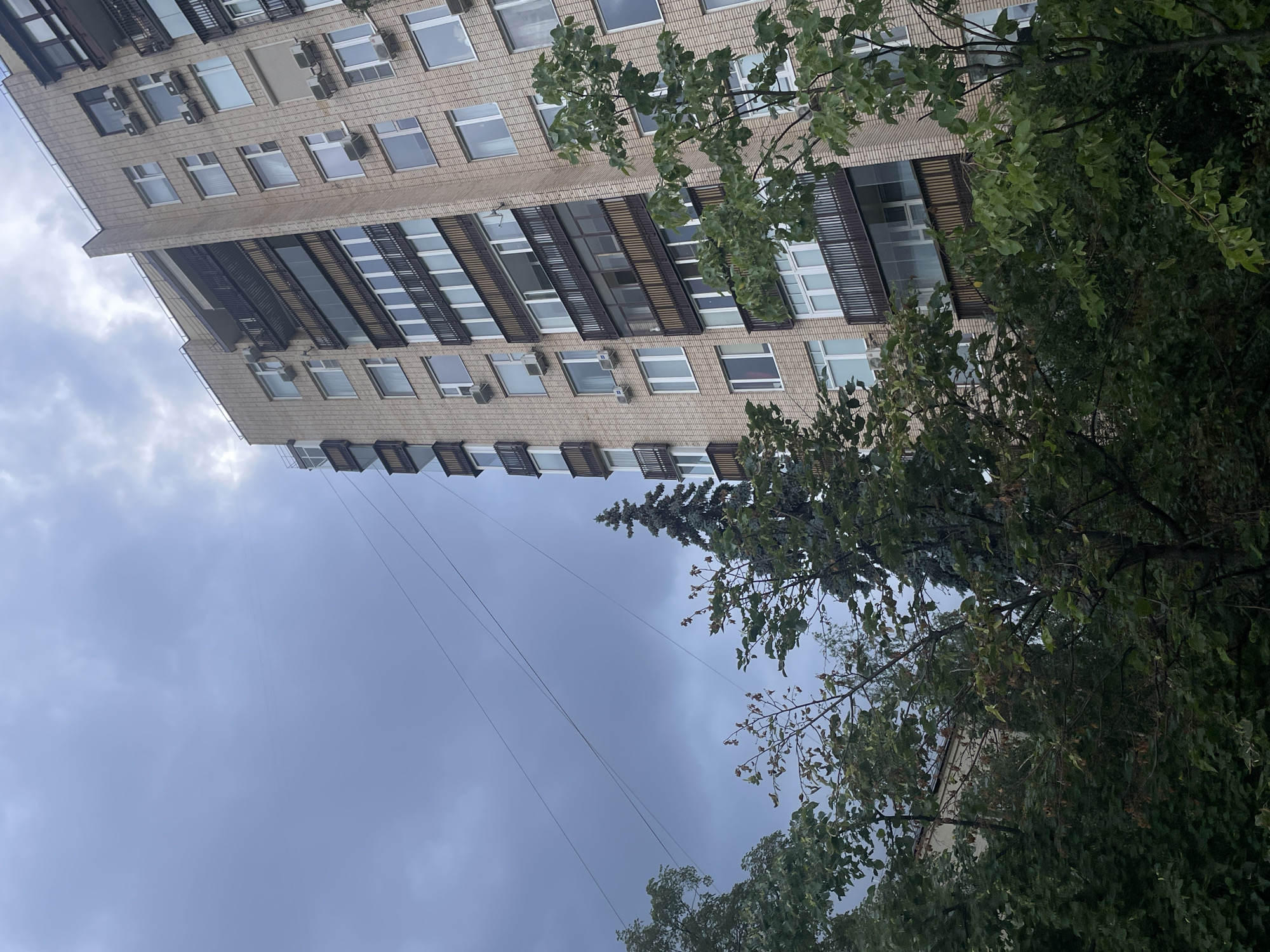 An exterior, low-angle, full shot depicts a multi-story apartment building in Moscow, Russia. The building's facade is clad in light-colored brick and features numerous balconies with dark railings. Air conditioning units are mounted on the exterior walls. The sky above is overcast with dark, heavy clouds. In the foreground, lush green trees partially obscure the view of the building and the sky. Several wires are visible crisscrossing the sky. The overall scene suggests a cloudy or possibly rainy day in an urban environment.