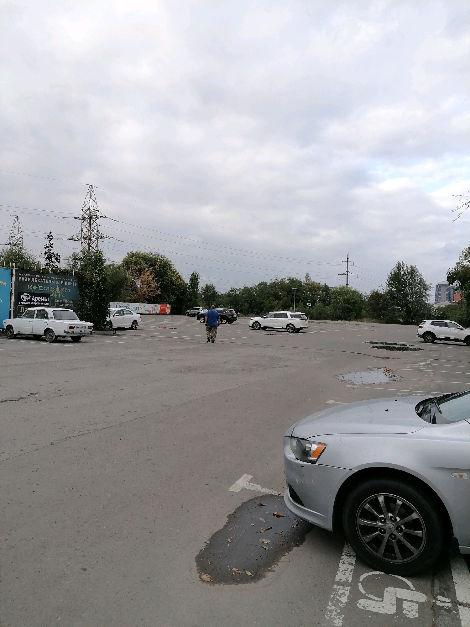 A man in a blue t-shirt and camouflage pants walks across a paved parking lot in Rostov-on-Don, Russia. The lot is mostly empty, with a few cars parked, including a white sedan on the left and two white SUVs further back. A silver car is parked in the foreground on the right, its front end in view. In the background, there are trees and tall power line towers. The sky is overcast. A banner on a fence in the background advertises a "Recreational Center 'Cosmos'" and "Arens Virtual Reality Attractions."