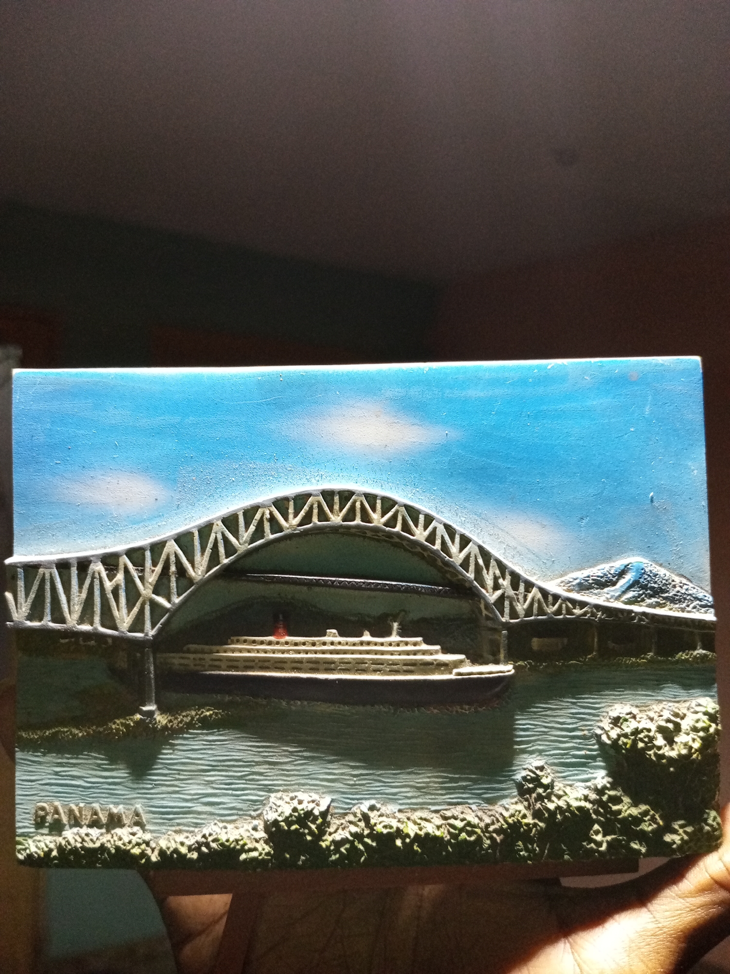 Here is a detailed description of the image:
The image shows a rectangular souvenir depicting the Panama Canal. A large passenger ship is sailing underneath a bridge. The bridge has a distinctive arched structure. The waterway is rendered with horizontal lines to simulate ripples on the water's surface. Green foliage is depicted along the waterfront. The sky is blue with a few white clouds. The word "PANAMA" is printed at the bottom of the souvenir.