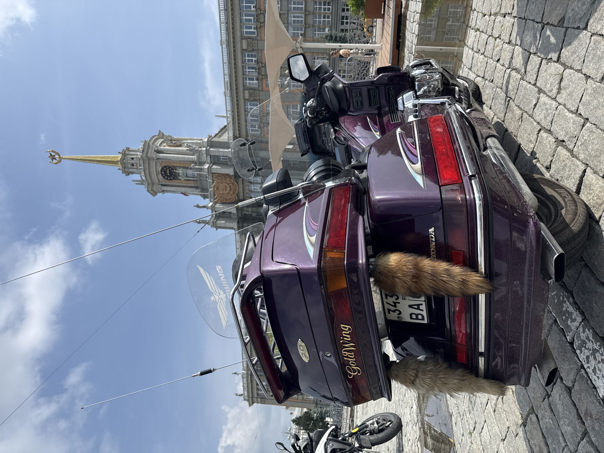 A metallic purple Honda Gold Wing touring motorcycle is prominently featured, parked on a cobblestone surface and angled slightly to the right, presenting its rear and right side to the viewer. The motorcycle is adorned with two vertical antennae, a clear windscreen featuring a white "WING" logo with an eagle graphic, gold "GoldWing" script on the rear top case, and a "40th Anniversary" badge. Below the partially visible Russian license plate ("343 RU BA"), two light brown fur tails hang. Dual exhaust pipes are visible at the rear. Reflections of the surrounding architecture and sky are visible in puddles on the cobblestones.
In the background, a large, ornate architectural building rises under a partly cloudy blue sky. This is the Sevastyanov House, located in Yekaterinburg, Russia. The building is characterized by its light-colored stone facade, numerous classical columns, decorative sculptures, and a prominent central clock tower with several clock faces. The tower culminates in a tall, golden spire topped with a large, golden five-pointed star ornament. Flags are visible near the clock tower. To the right of the motorcycle, a person in light-colored clothing stands near an outdoor seating area with distinctive white, sail-like sunshades. The front wheel and suspension of another motorcycle are partially visible on the far left.