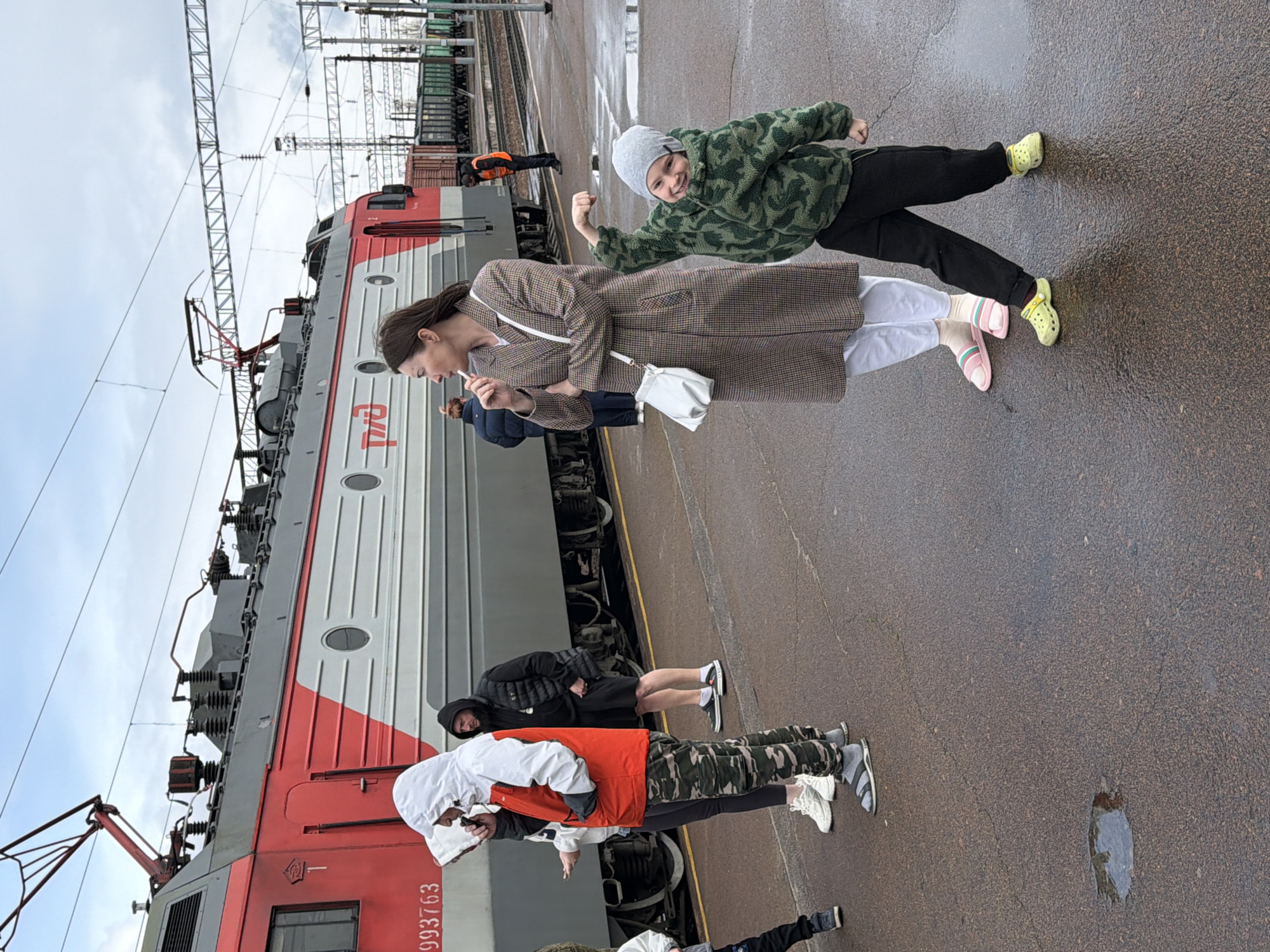 A group of people are standing on a train platform next to a red and grey electric locomotive in Liski, Russia. The locomotive has a white "RZD" (Russian Railways) logo on its side and the number "3ЭС5К-993763" painted on its front. Overhead are electrical wires and supporting structures. One person is standing in front, facing the locomotive and wearing an orange vest and white hooded sweatshirt. A person behind them wears a black hooded jacket and black shorts, and stands next to the locomotive wheels. A woman in a plaid coat holds something to her mouth and has a white purse slung over her shoulder. Next to her, a younger person in a green patterned coat and grey knit hat holds a fist in the air, possibly posing for the camera. The ground is wet and reflective, suggesting it has recently rained.