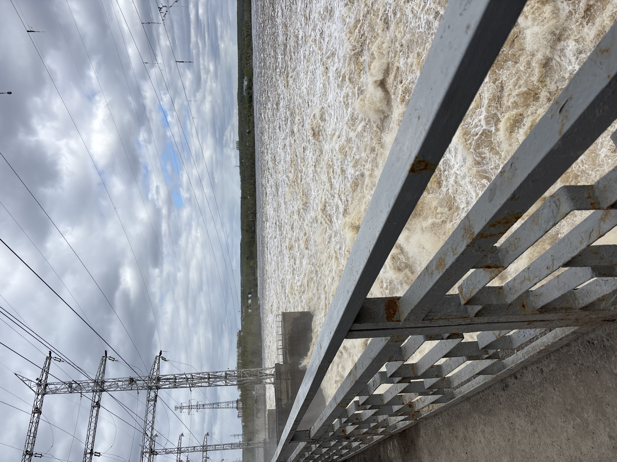 The image shows a high-angle view of water cascading over a dam in Perm, Russia. A gray metal railing occupies the foreground on the right, and power lines and utility towers are visible on the left. The water is turbulent and brown, suggesting a high volume of flow, possibly due to recent heavy rainfall or snowmelt. A strip of trees lines the distant shoreline. The sky is overcast with a mix of gray and white clouds.