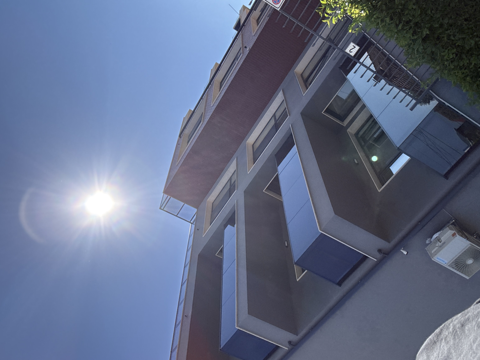 The image shows a low-angle shot of a modern building with a dark gray facade, blue-tinted balconies, and red brick accents. The sun is bright in a clear blue sky, creating a lens flare. On the ground level, an outdoor air conditioning unit is visible, along with a gray textile. A metal railing with a sign displaying the number "2" is present on the right side of the frame, partially obscured by green foliage. The architectural style suggests an urban setting, possibly in Varna, Bulgaria, given the context. The perspective is tilted, giving a dynamic feel to the composition.