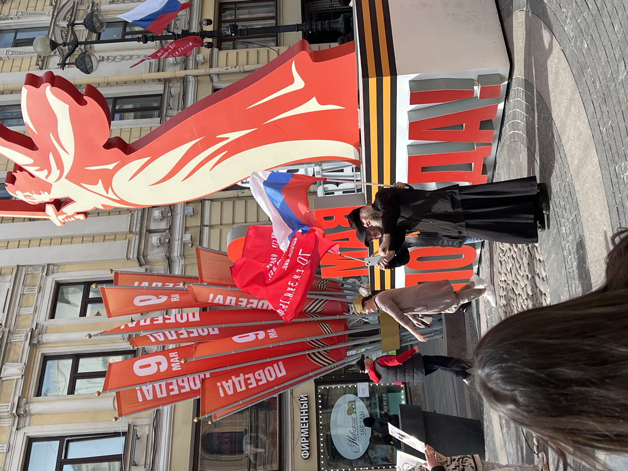 The image shows a street scene in Saint Petersburg, Russia. Banners and flags are displayed, indicating a celebration or commemoration. The predominant color scheme is red, white, and orange, typical of Russian patriotic displays. Flags with the inscription "9 МАЯ ПОБЕДА!" (May 9 Victory!) are visible, referencing Victory Day, a Russian holiday celebrating the victory over Nazi Germany in World War II. There is a large structure with the words "Днём Победы!" (Victory Day!). A person in a black robe is holding a Russian flag. Several other people are present, observing or involved in the event. The architecture of the surrounding buildings is characteristic of central Saint Petersburg. A shop sign reads "ФИРМЕННЫЙ НЕВСКИЕ ЗВЕЗДЫ" (branded Neva stars). There is also an orange and black striped St. George ribbon on display.