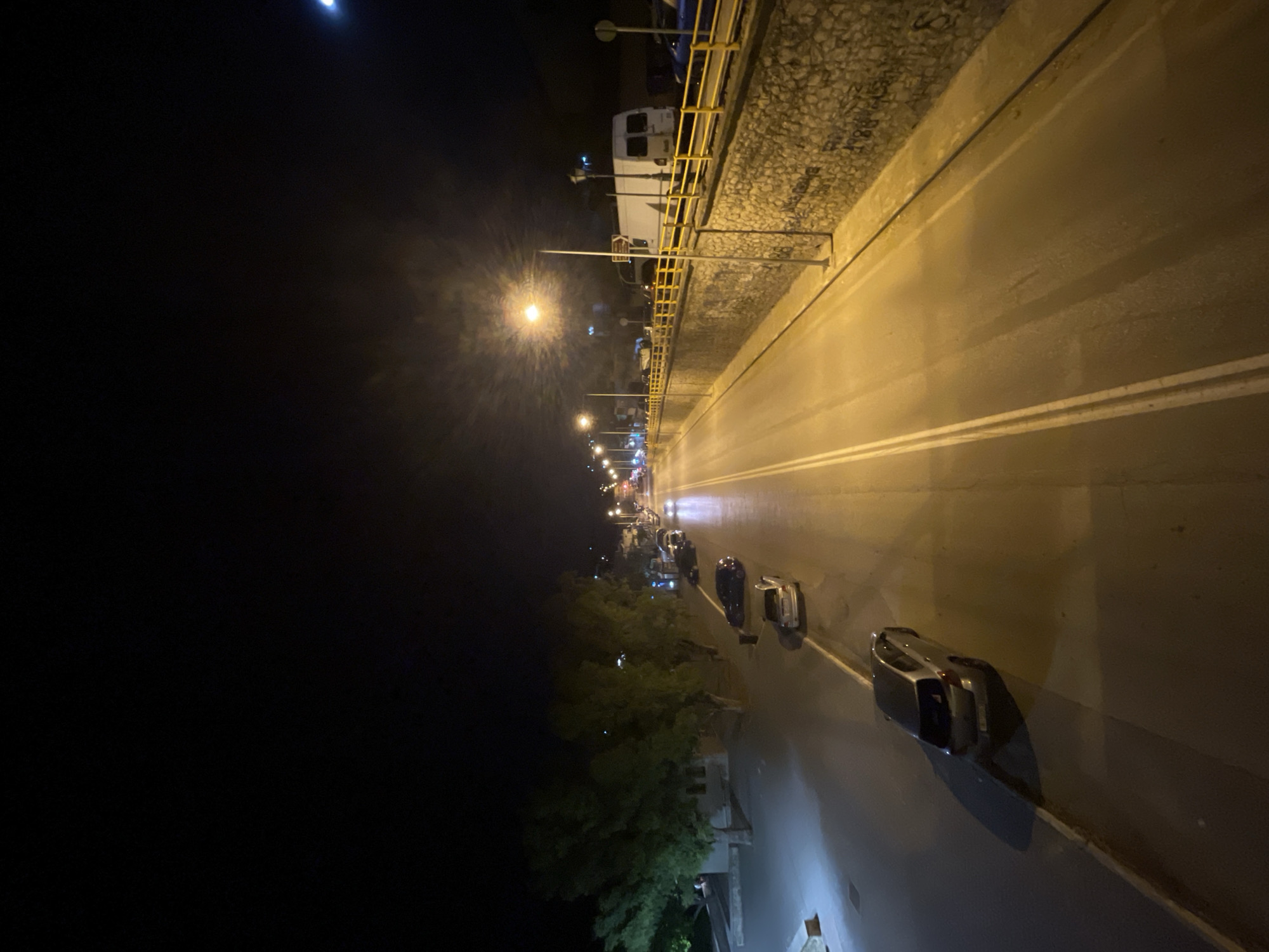 The image depicts a nighttime view of a street in Kassandra Municipal Unit, Greece. The street is illuminated by streetlights, casting a warm glow on the asphalt and surrounding areas. Several cars are parked along the right side of the road, and a few are driving on it. A white van is visible parked on the right side, beyond the yellow barrier. The street has double white lines marking the lanes. A stone wall with graffiti runs along the right side of the road, topped with a yellow railing. Trees with dense foliage are present on the left side of the street, with some buildings visible behind them. The sky is dark with no stars visible, but a faint glow from a distant light source is present in the upper right corner. The overall scene suggests a quiet urban or semi-urban environment at night.