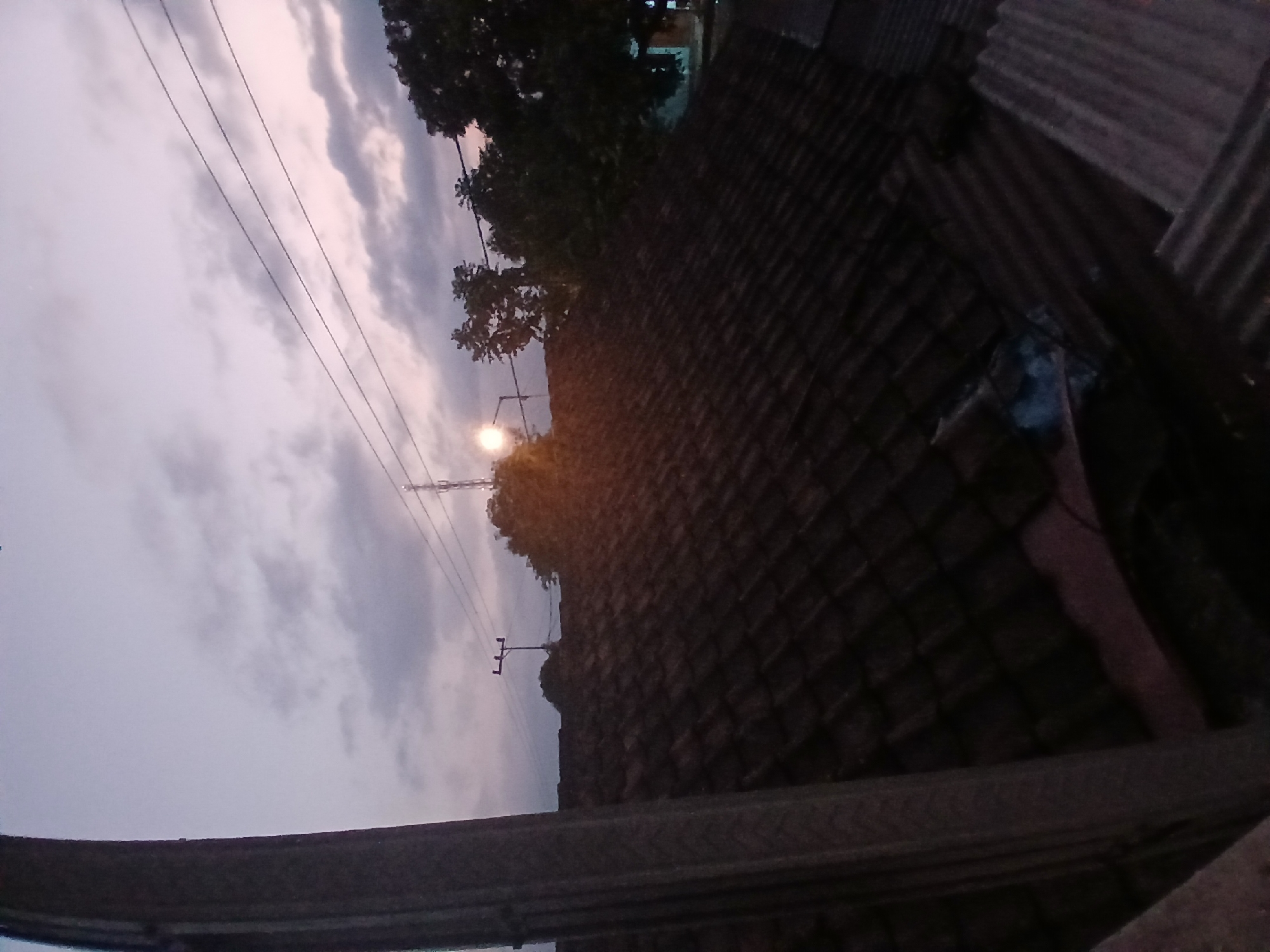 An outdoor, medium shot captures a twilight sky above a tiled rooftop in Tenggarang, Indonesia. The sky is a gradient of purple and pink hues, with soft clouds scattered across it. Three dark, thin power lines stretch horizontally across the upper portion of the frame. In the distance, silhouetted trees and a tall transmission tower are visible against the fading light. A single streetlamp casts a bright, yellow glow near the left side of the image. The foreground is dominated by the dark, textured surface of a tiled roof, which occupies the majority of the lower half of the image. The roof appears weathered, with individual tiles clearly distinguishable. The overall atmosphere is calm and serene, characteristic of the transition from day to night.
