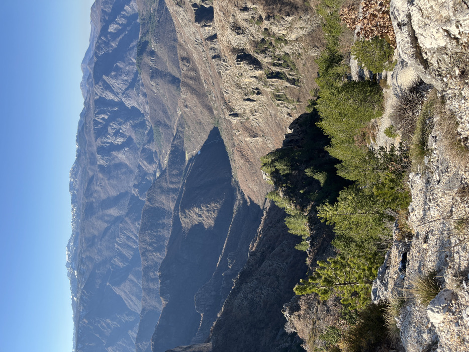 This image depicts a vast mountainous landscape in Klobuk, Bosnia and Herzegovina, under a clear blue sky. The foreground consists of rough, rocky terrain sparsely covered with light-colored dry grasses and small, scattered coniferous trees. A dark, irregular object, possibly a discarded item, is partially visible on the ground among the rocks in the lower-left quadrant.
The midground reveals a series of deep valleys and ridges, characterized by steep slopes of exposed rock and areas covered with a mix of bare deciduous trees and clusters of evergreen foliage. Distinct shadows fall across these geographical features, indicating direct sunlight from an elevated position.
In the background, multiple layers of mountains extend to the horizon, gradually fading in color intensity. The most distant peaks are capped with visible patches of snow. A lighter-colored strip, consistent with a road or cleared pathway, is discernable within a valley in the mid-to-far distance. No people, animals, or structures are prominently featured in the scene.