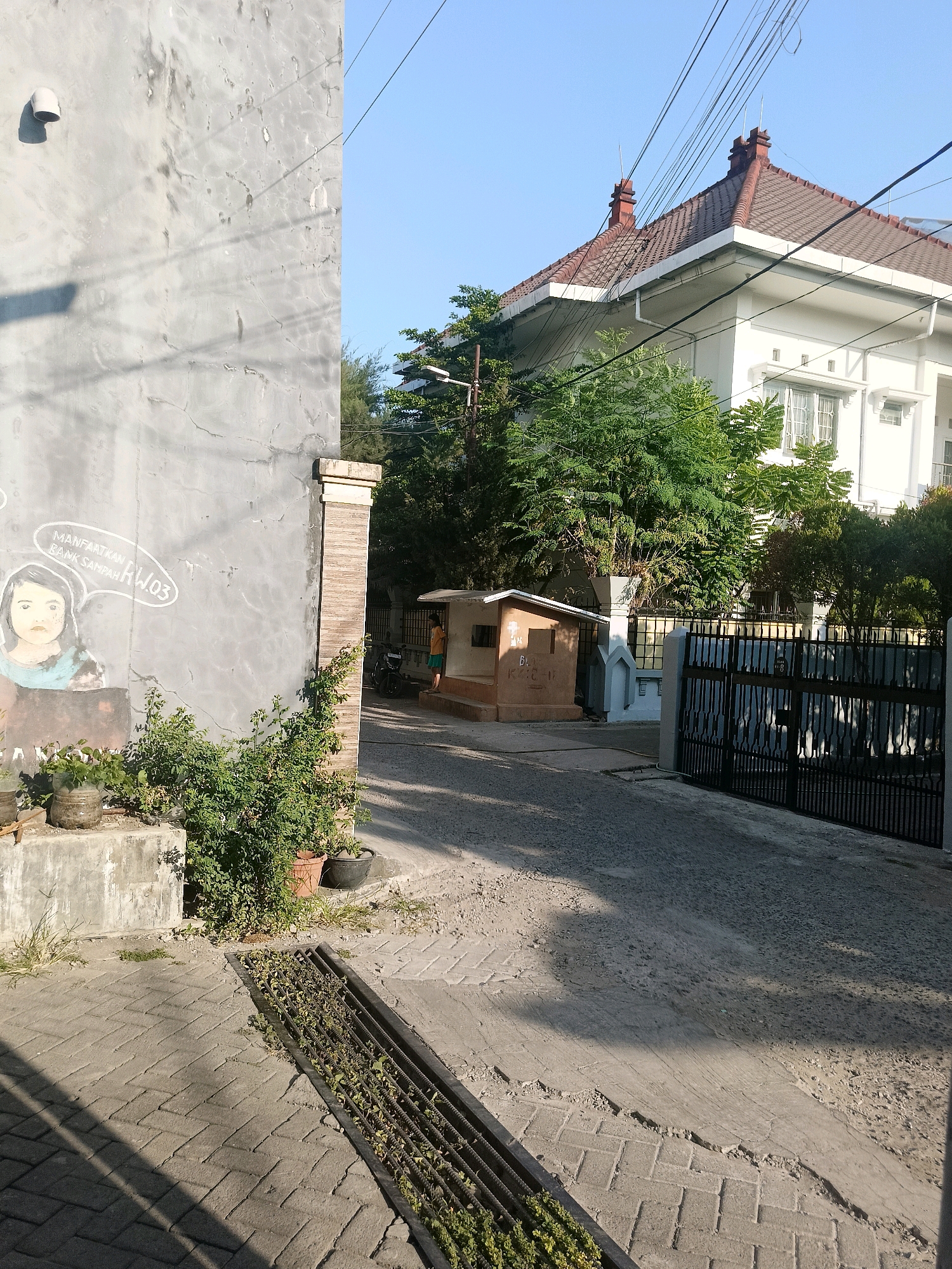 The image depicts a residential street scene in Makassar, Indonesia. On the left, a rough, grey concrete wall with a painted mural of a young girl's face and text in Indonesian is visible. Above the mural, a white pipe protrudes from the wall. Adjacent to the wall is a brick pillar. In the center of the image, a small, brown, shed-like structure with a white roof and a small window stands on a raised concrete base. A person wearing a yellow shirt and orange shorts is standing near this structure. To the right, a large white house with a red tile roof and multiple windows is partially visible, surrounded by lush green trees. In front of the house, a black metal gate stands closed. The street is paved with concrete and brick pavers, with a metal grate for drainage in the foreground. Power lines are strung across the clear blue sky. The lighting suggests it is daytime, likely morning or afternoon, with distinct shadows cast across the ground.