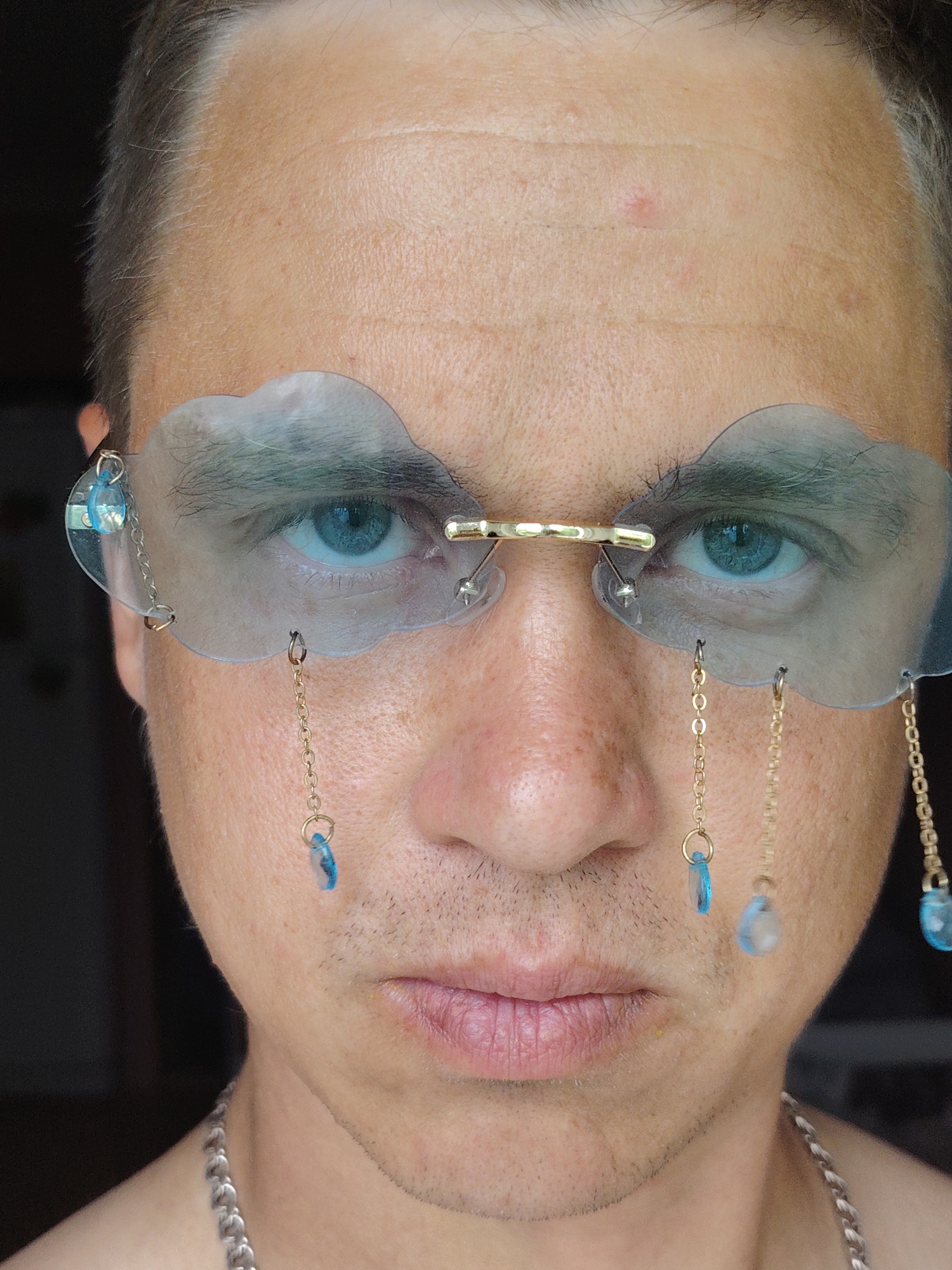 A close-up, frontal portrait depicts an adult male with fair skin and light blue eyes, looking directly at the viewer. His short, light-brown hair is visible at the temples, and faint stubble covers his lower face and chin. His forehead exhibits several horizontal lines and a few small red blemishes. He wears unique, rimless glasses featuring two clear, cloud-shaped lenses connected by a small gold-colored bridge piece across the nose. From various points on the lenses, thin gold-colored chains descend. On the viewer's left, one chain from the upper outer edge carries a small blue teardrop bead, connecting to another small blue bead below. On the viewer's right, three separate chains with small blue teardrop or oval-shaped beads dangle from the lower edge of the lens. A chunky, silver-colored chain necklace is visible around his neck. The background is uniformly dark and out of focus, providing no discernible environmental details. The location is not identifiable from the image.