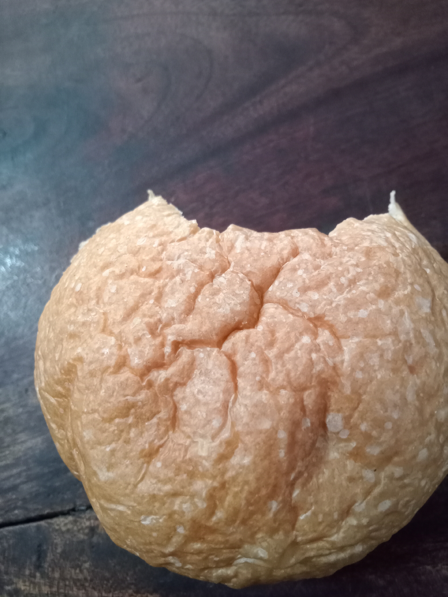 A close-up, eye-level, vertical shot shows a round, light brown bread roll on a dark brown wooden surface. The bread roll appears to have been bitten into, with a crescent-shaped portion missing from the top. The surface of the bread is textured with a series of irregular indentations and lighter colored flecks, possibly flour. The wooden surface displays visible grain patterns and a slightly worn appearance. The lighting is dim, creating shadows and highlighting the texture of the bread and wood. The scene is devoid of any people or specific landmarks that would indicate a location within Simo, Indonesia.