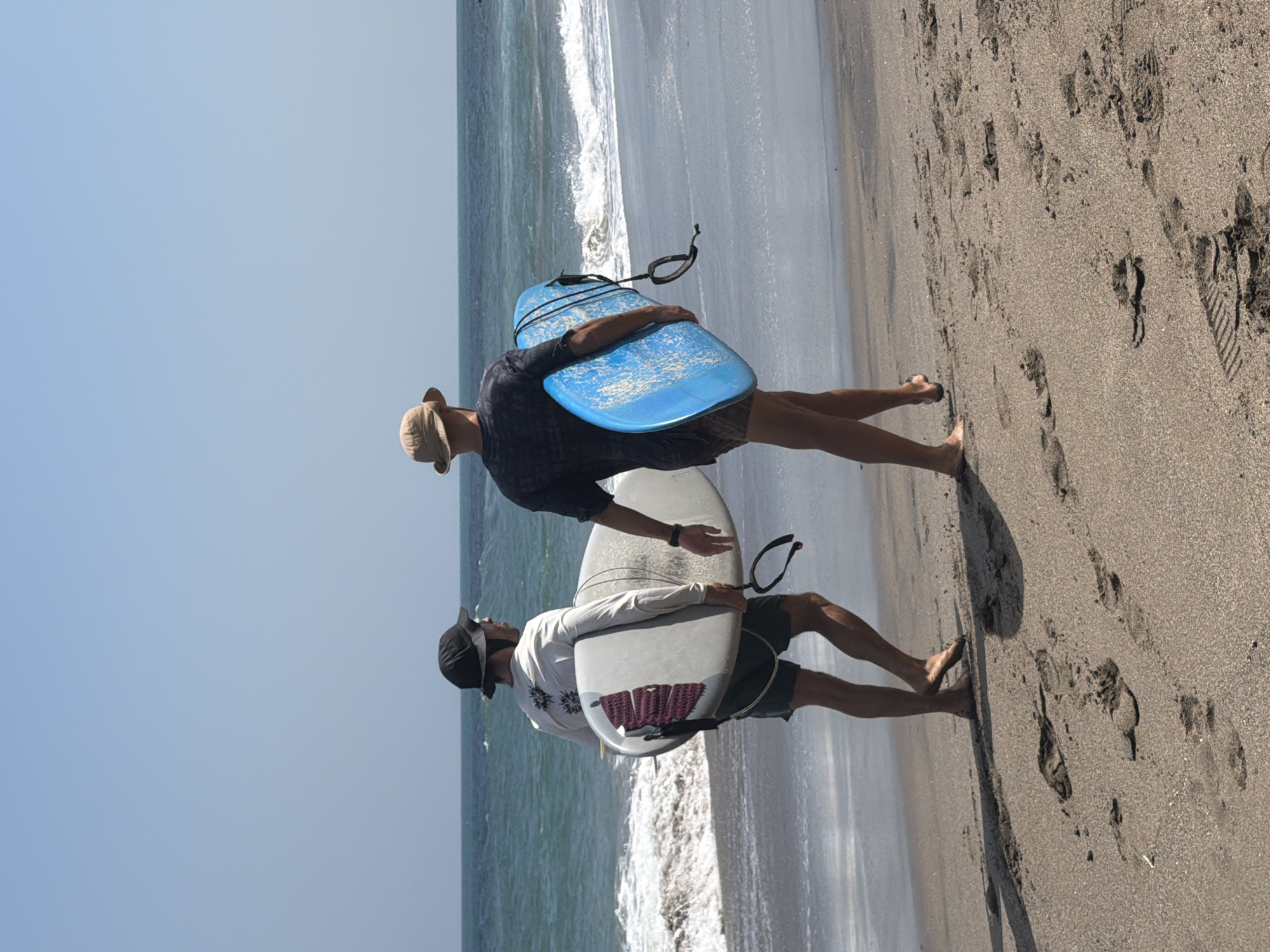 Two men are walking on a sandy beach in North Kuta, Indonesia, carrying surfboards. The man on the left is wearing a black bucket hat, a long-sleeved white shirt with a graphic print, and dark shorts. He is carrying a white surfboard with a pink grip pad. The man on the right is wearing a light brown bucket hat and a dark patterned t-shirt with dark shorts. He is carrying a bright blue surfboard. Both men are walking away from the camera towards the ocean, which is visible in the background with waves. The sky is clear and blue. Footprints are visible in the sand.