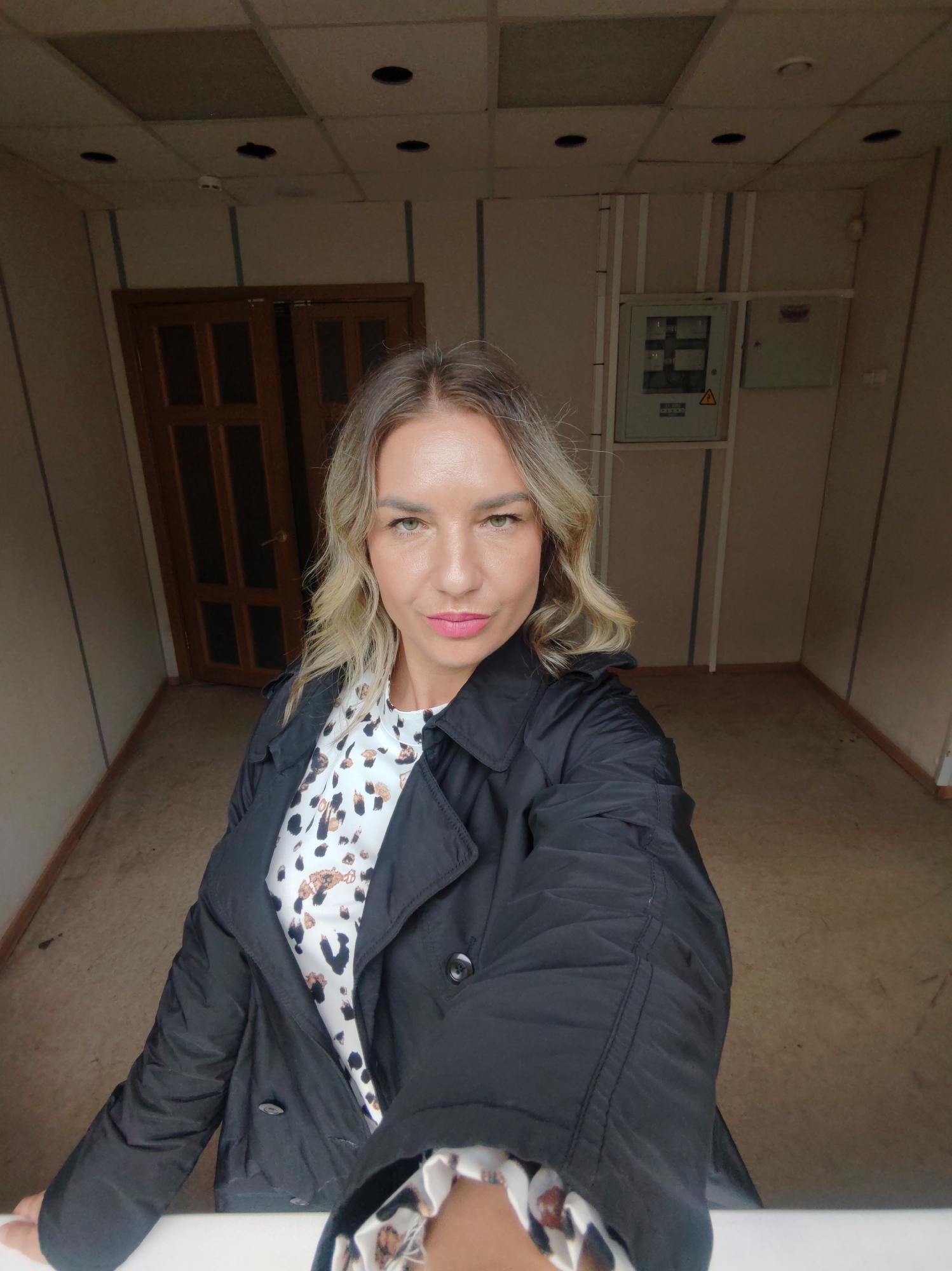 A woman stands in an empty room. The walls are beige with vertical stripes. The ceiling is a drop ceiling with recessed lights. There is a wooden door in the background to the left and electrical panels on the right. The floor is beige. The woman has blonde hair and is wearing a black coat over a patterned top. She is looking at the camera. The location is Unknown, Unknown.