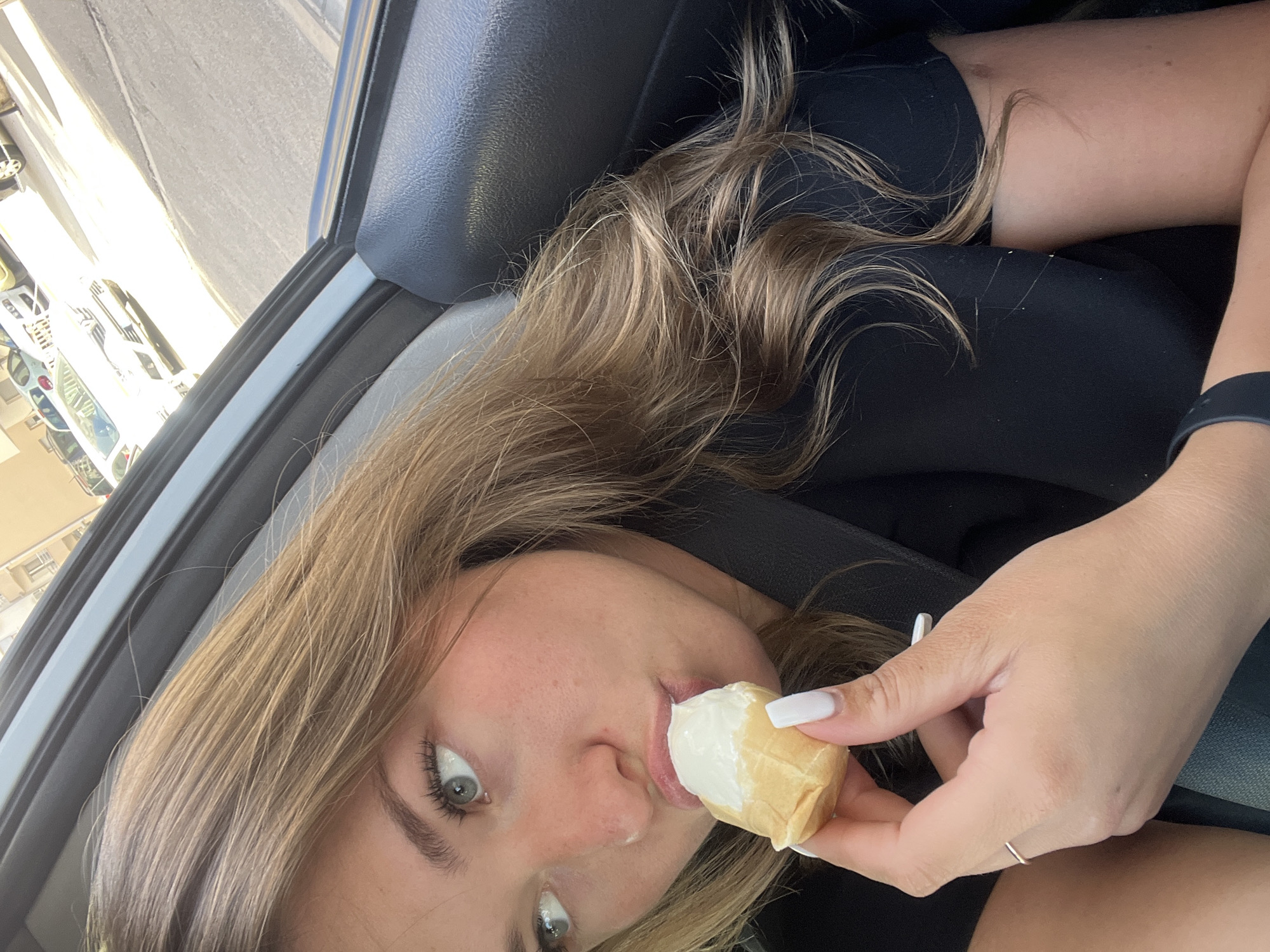 A young woman with blonde hair and light eyes is eating an ice cream cone in a car. She is wearing a black top and has white nail polish. The car is parked in a street in Budva, Montenegro. Other cars and buildings are visible in the background.