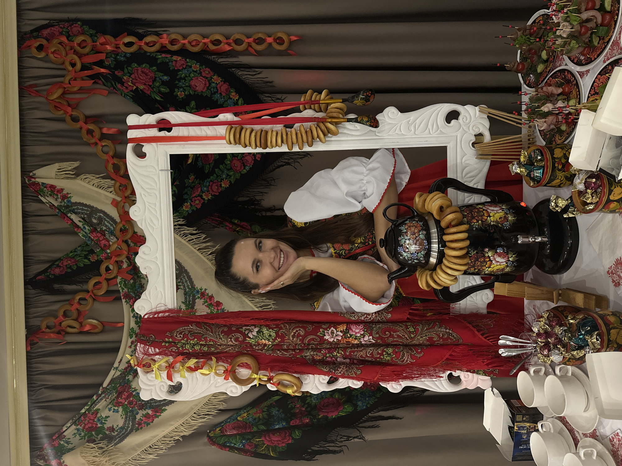 A woman is posing in front of a white frame decorated with a samovar, bagels, and traditional Russian shawls. She is wearing a traditional Russian dress with a white blouse and red and white trim. She has her hand to her face and is smiling. The background is a grey curtain. A table with cups, plates, and food is to the right of the frame. The location is unknown.