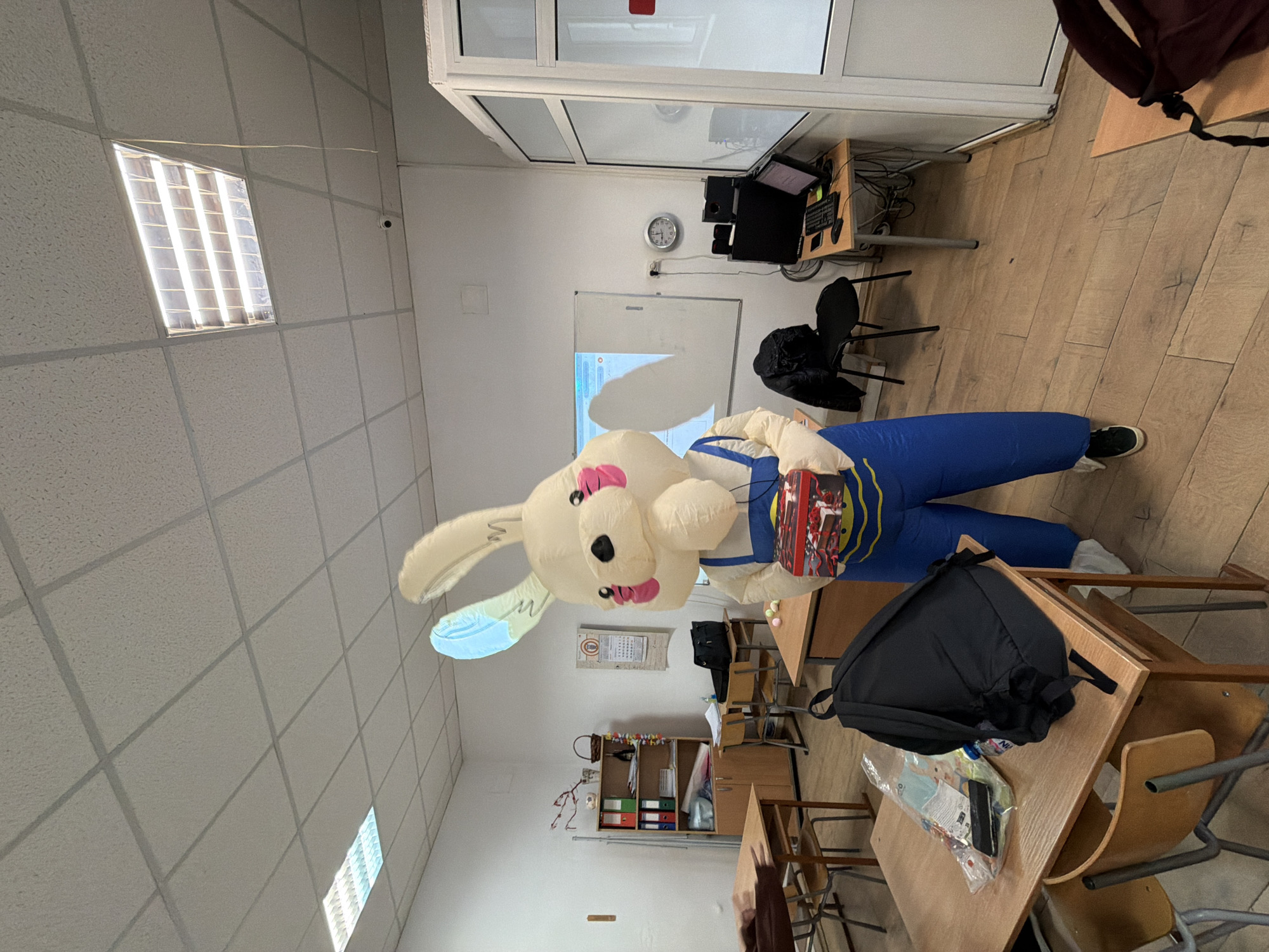 An interior view of a room in Panagyurishte, Bulgaria, depicts an inflatable bunny figure wearing blue overalls, situated in a classroom setting. The bunny is carrying a box, possibly a toy. Several rows of desks and chairs fill the room, with backpacks resting on some of the desks. A whiteboard is visible on one wall, and computer equipment is visible on a nearby desk. The floor appears to be made of wood, and the ceiling is a standard tile grid with fluorescent lighting.