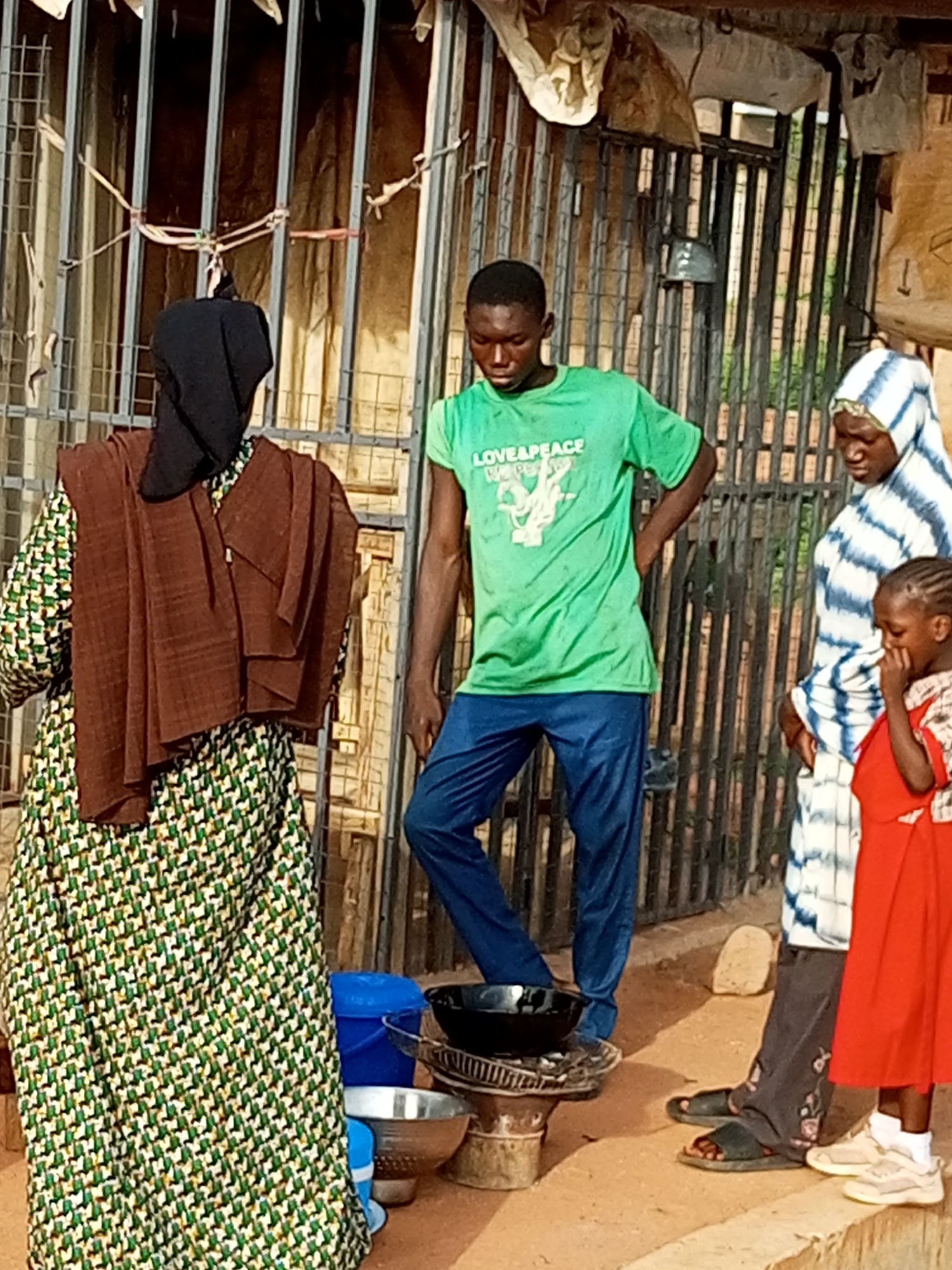 In Tumfure, Nigeria, a young man in a green "LOVE&PEACE" t-shirt and blue pants stands with his hands on his hips. To his right, a woman wearing a blue and white striped hijab and a woman wearing a brown hijab and a patterned green dress are also present. A young girl in a red dress stands next to the woman in the hijab. In front of the man, a cooking stove with a black pot on top is visible. Two metal bowls and a blue bucket are also on the ground. The scene takes place outdoors with a metal fence in the background.