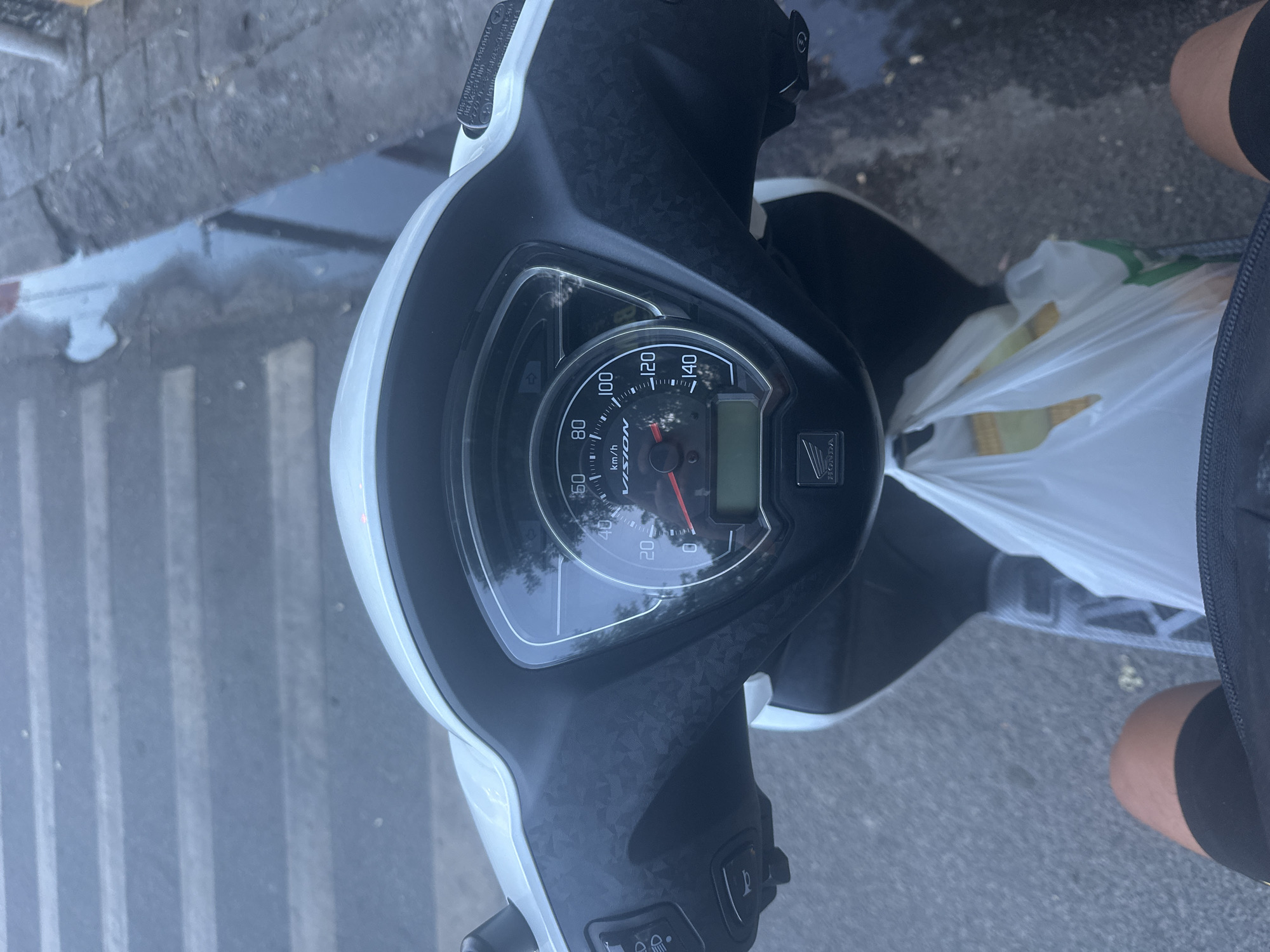 A first-person perspective shot shows a rider on a Honda Vision scooter in Nam Nha Trang Ward, Vietnam, at eye level with the handlebars and dashboard. The scooter's instrument cluster is visible, with the needle on the speedometer pointing to approximately 20 km/h. A digital display is below the analog speedometer. A white plastic bag, presumably containing groceries, is placed on the floorboard of the scooter. The rider's legs are visible in the foreground, wearing black shorts. The road surface appears wet, with reflections indicating recent rain or puddles, and a pedestrian crossing is visible in the upper left of the frame. The environment is urban, with hints of trees and buildings in the reflection on the scooter's instrument panel.