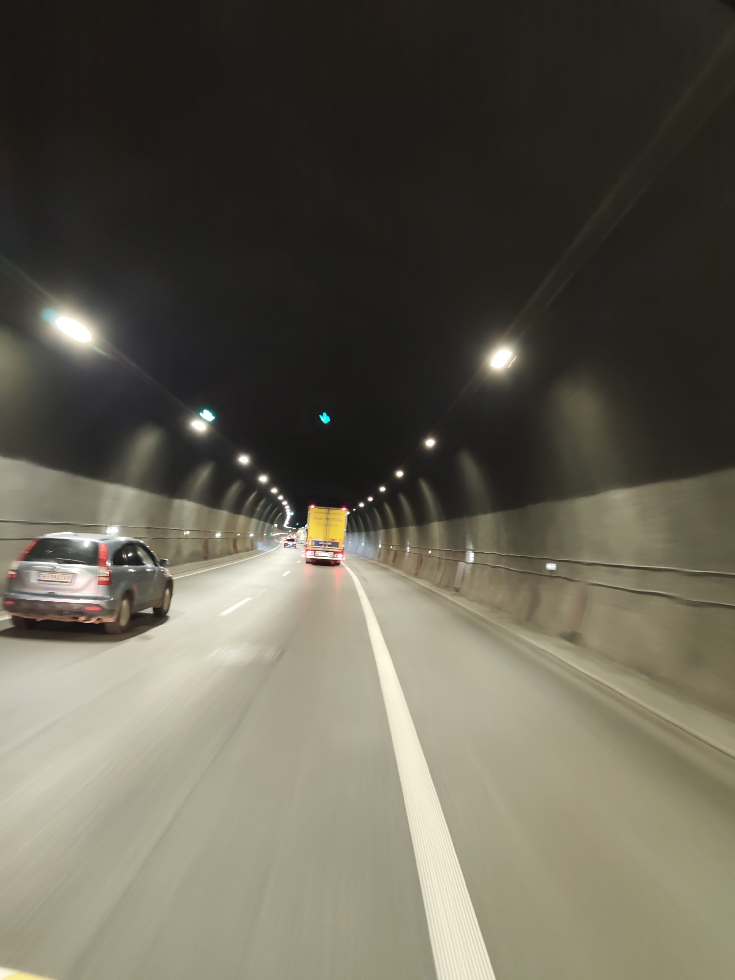 The image captures a forward-looking perspective from within a highway tunnel. The tunnel is illuminated by rows of lights mounted on the ceiling, casting a bright glow on the roadway. The walls of the tunnel appear to be made of concrete. Several vehicles are visible on the road: a silver SUV is in the left lane, and further ahead, a yellow truck with its brake lights illuminated is in the right lane. The road is marked with a white line separating the lanes, and there are some traffic lights visible, showing green signals. The overall impression is one of movement and transit through an enclosed passage. The location is identified as being in the city of Gorno Kamartsi, Bulgaria.