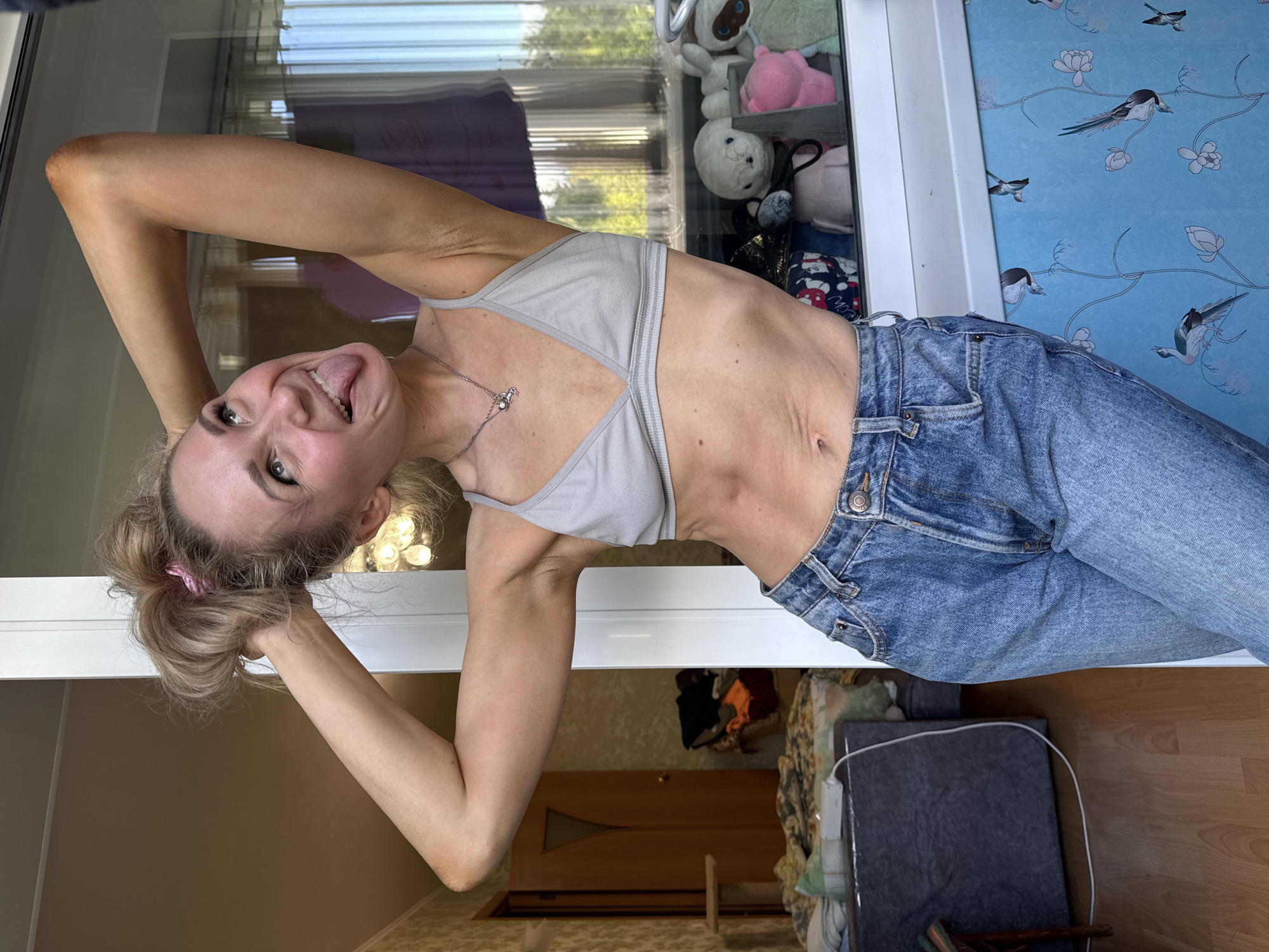 This media file captures a full-body shot of a young woman in a playful pose, leaning backward with her head tilted and tongue sticking out. She is wearing a light grey sports bra and blue denim jeans. Her hair is pulled back in a messy bun with a pink scrunchie. The background appears to be an indoor setting, possibly a room in a house. A large window or glass door dominates the left side of the frame, reflecting some light and showing a blurred interior on the other side. On the right side, a wall is decorated with a blue wallpaper featuring a bird and floral pattern. Several stuffed animals are visible on a shelf or ledge behind the woman, partially obscured by the reflection and her pose. The overall lighting suggests it's daytime, with natural light coming from the window. The woman's expression conveys a sense of fun and lightheartedness. The location context provided is Perm, Russia.