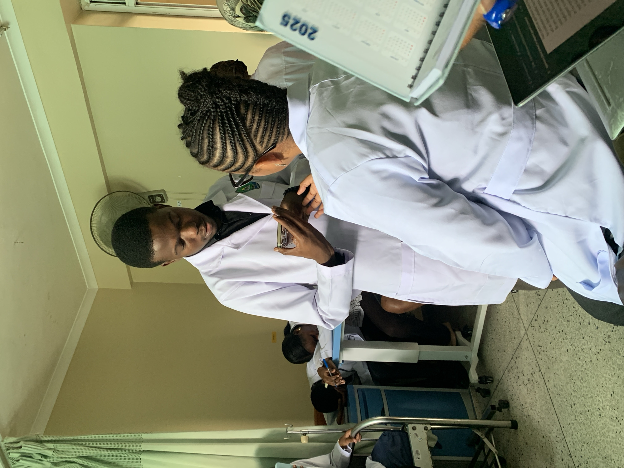 The image depicts an indoor scene, identified as a medical training or clinical environment in Dar es-Salaam, Tanzania.
In the foreground on the right, the back of a person, likely female, with intricate cornrow braids and glasses, is visible. This individual wears a light-colored medical uniform or lab coat and holds a light green spiral-bound calendar marked "2025" in their right hand, with a dark electronic tablet partially visible beneath it. Facing this person in the center of the frame is a male individual, wearing a white lab coat over a dark collared shirt and glasses. He holds a smartphone horizontally in both hands, appearing to display its screen to the person in the foreground.
In the mid-ground on the left, a person in a white lab coat is partially visible, seated next to a metallic IV pole on wheels. Further into the background, two individuals are seated at a light blue table or desk. One female, wearing a white lab coat, is resting her head down on the desk, with a pen visible in her left hand. Another person is partially visible seated behind her.
The room features light tan walls with white crown molding. A wall-mounted oscillating fan is visible on the right wall, along with a partial window frame. Green and white striped curtains are drawn to the left. The ceiling is off-white with some minor discoloration. The floor is composed of light-colored, speckled terrazzo or tile.