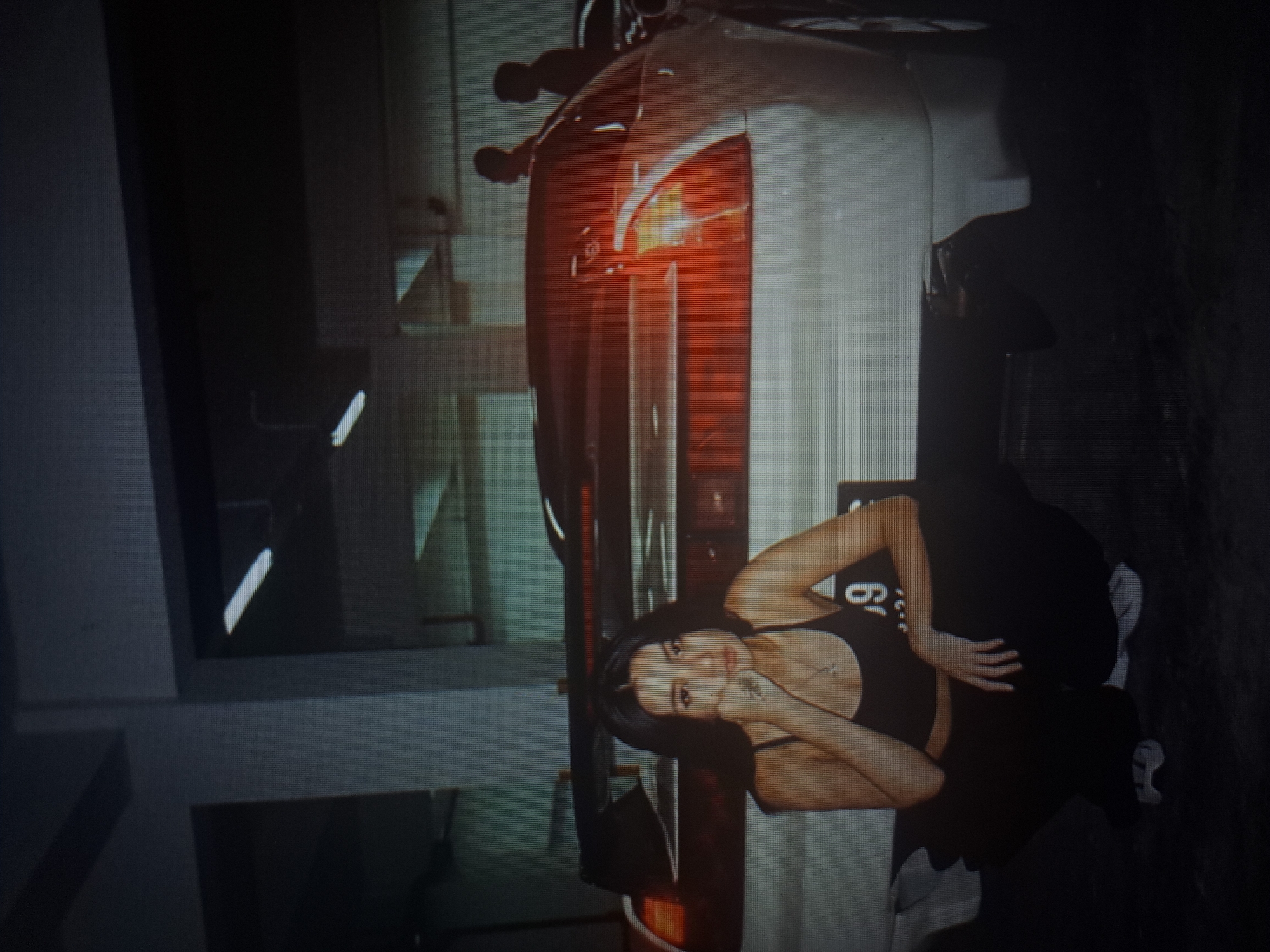 A medium shot depicts a woman posing next to a parked white car in what appears to be an indoor parking garage in Yerevan, Armenia. The woman, with dark hair, is wearing a black tank top and black pants. She is partially leaning against the car, with her left hand near her face and her right hand resting on her left hip. The car, a sleek, modern model, dominates the center of the image. The background shows elements of a typical parking garage, including overhead lighting fixtures and concrete structures. The light is somewhat dim, adding a slightly dramatic atmosphere to the image.