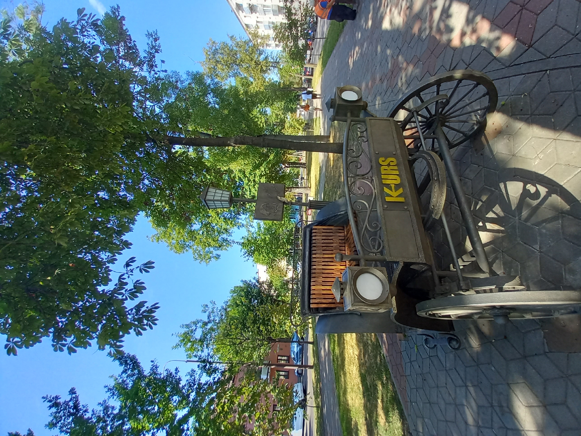 The image shows a sunny day in Brest, Belarus, with a view of a decorative horse-drawn carriage monument. The monument, made of metal with wooden seats, is placed on a paved sidewalk lined with trees. In the background, there are residential buildings and cars on the street. Several people are walking on the sidewalk. The monument has the inscription "K-URS" in yellow letters.