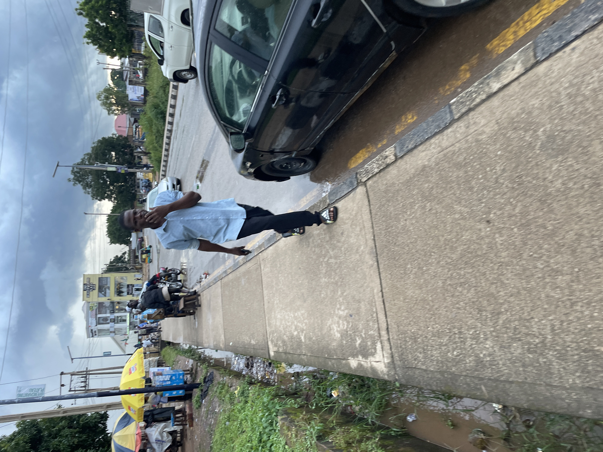 A medium shot captures a man walking on a sidewalk adjacent to a road in Jos, Nigeria. The man, wearing a light blue button-down shirt and black pants, walks towards the camera, touching his chin with his left hand. The sidewalk is made of concrete slabs, with a drainage ditch to its left filled with water and debris. Beyond the ditch, a few small market stalls with umbrellas and various goods are visible. The road is wet, indicating recent rain, with a black car parked on the right side of the frame. In the background, buildings and trees line the street, under a cloudy sky.
