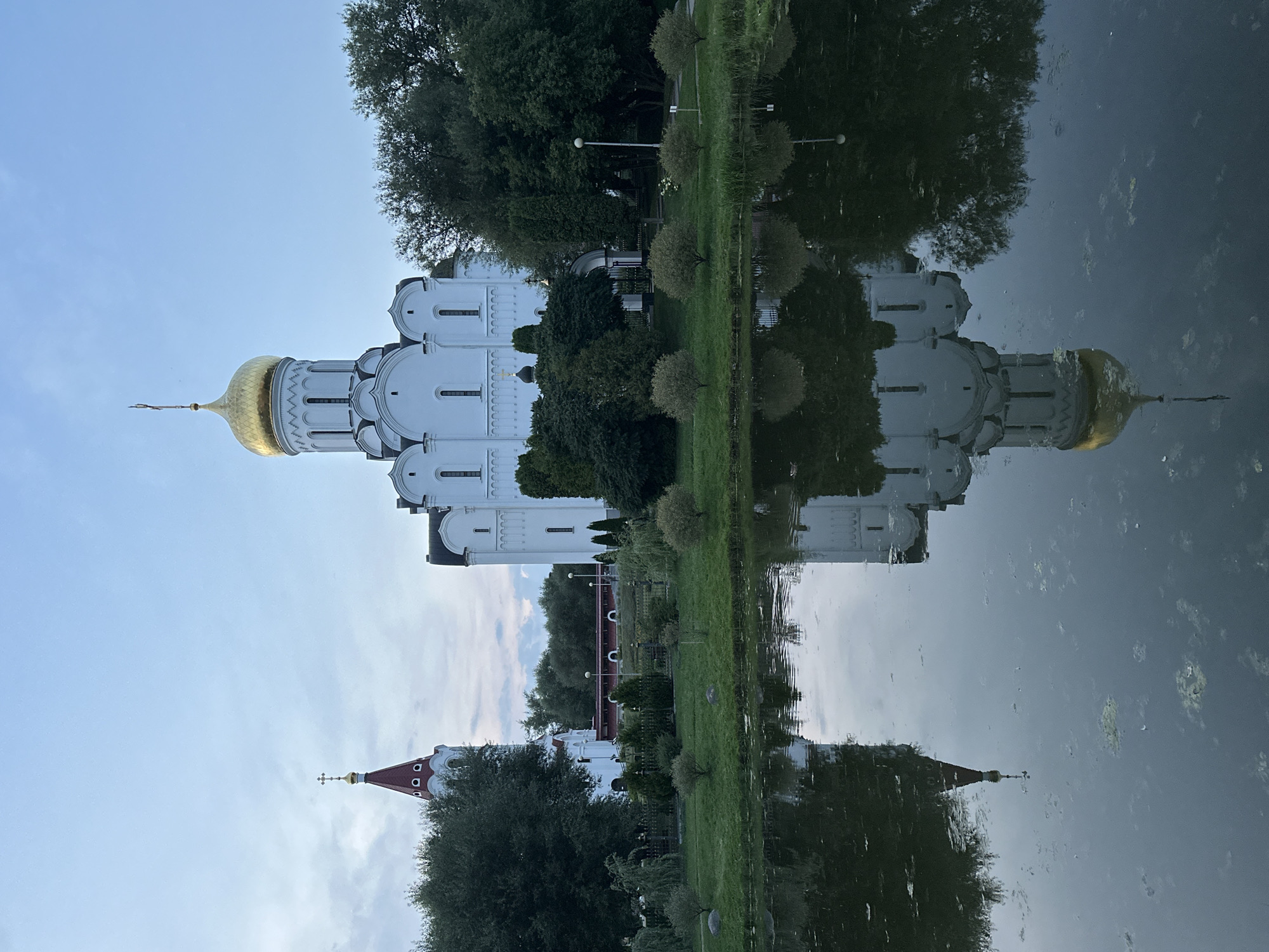 A white Orthodox church with a golden dome is reflected in a pond in front of it. To the left of the church, a bell tower with a red roof and a golden cross is visible. Trees and a manicured lawn surround the church and pond. The sky is overcast with a few clouds. The scene appears to be captured at dusk or dawn, with soft, diffused light. The location is an outdoor environment, likely a park or religious complex in Unknown, Unknown.
