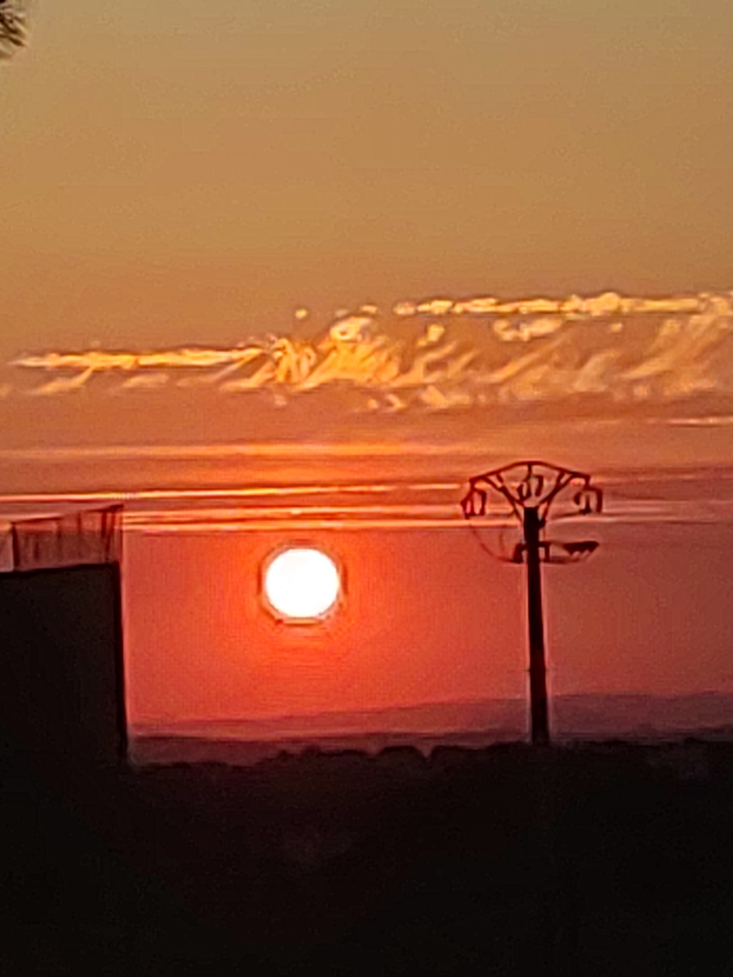 The image captures a sunset over a body of water, likely a reservoir, near Sobradillo, Spain. The sky is a gradient of warm oranges and yellows, with scattered clouds tinged with the same hues. In the foreground, a dark silhouette of a structure, possibly a building, occupies the left side of the frame. To the right of this structure, a circular window with a bright, luminous sun visible through it is centrally positioned against a vibrant red and orange background, suggesting the light is emanating from a source within or behind the window. Further to the right, a power pylon stands silhouetted against the sunset. The horizon line is blurred, with faint, dark shapes suggesting distant landforms. The overall impression is of a tranquil, natural scene illuminated by the warm glow of the setting sun, with man-made elements subtly integrated into the landscape.