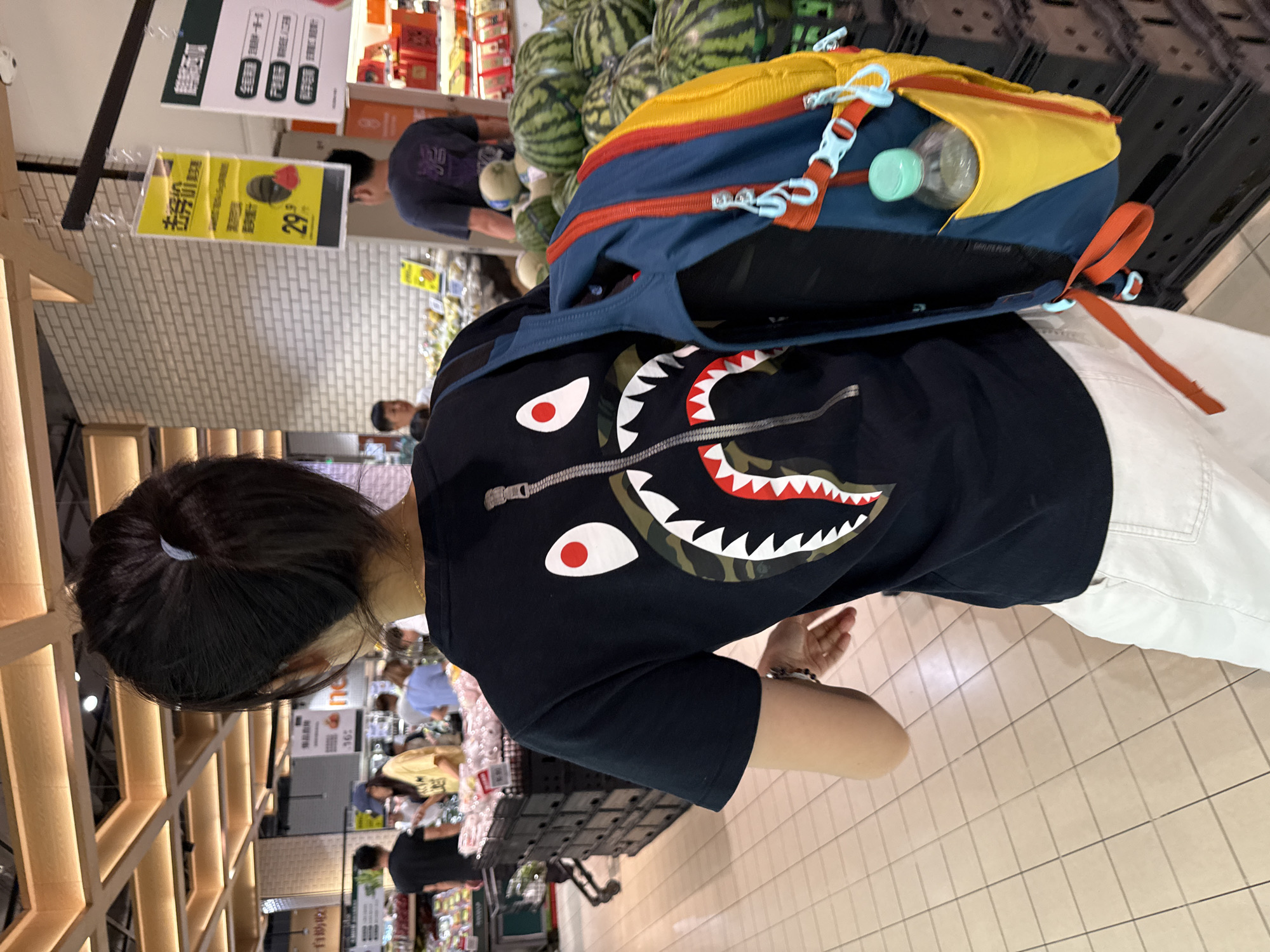 In a brightly lit supermarket aisle, a person from behind is wearing a black t-shirt with a shark graphic print and white pants. They are carrying a multi-colored backpack with a yellow side pocket containing a clear plastic water bottle. The person is walking past shelves stocked with produce, including watermelons. In the background, other shoppers are visible, and signage in Chinese is displayed on the walls. The floor is tiled in a beige color. The scene is located in the Haizhu District of China.