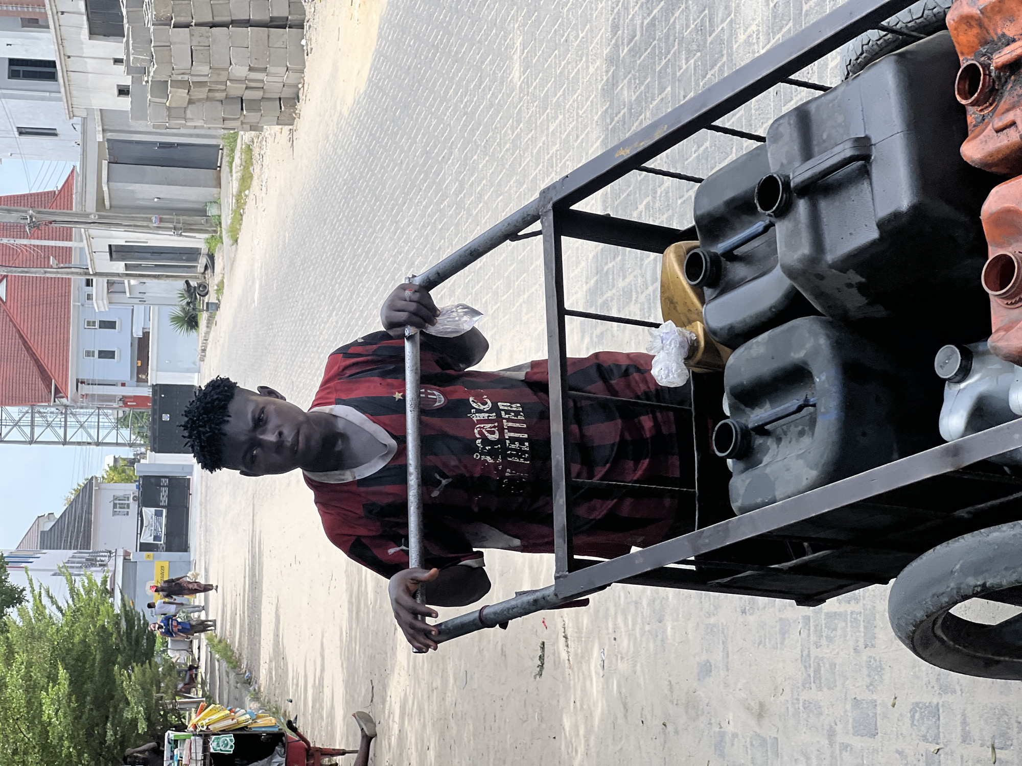 In an outdoor scene in Igboefon, Nigeria, a young man is prominently featured, holding onto a metal handcart or trolley filled with plastic containers. The man, with short dark hair and a neutral expression, looks directly at the camera. He wears a red and black striped jersey with "AC MILAN," "BITCOIN," and "FUTUREREADY" visible text on it.
The handcart, constructed from metal bars, carries several jerrycans of varying colors (black, orange, yellow), suggesting the transport of liquids like fuel or water. Some of the jerrycans appear worn. The man's hands grasp the horizontal bar of the cart.
Behind him, a paved street stretches into the distance, lined with buildings and some greenery. On the right side of the street, a large stack of concrete building blocks is visible. Further down the street, other individuals are seen walking, and a motorcycle is parked. The sky is bright, indicating it's daytime with clear weather. A distant sign features the text "YOU ARE WELCOME." The overall setting suggests a developing urban or semi-urban environment, possibly a residential or commercial street where goods are transported manually.