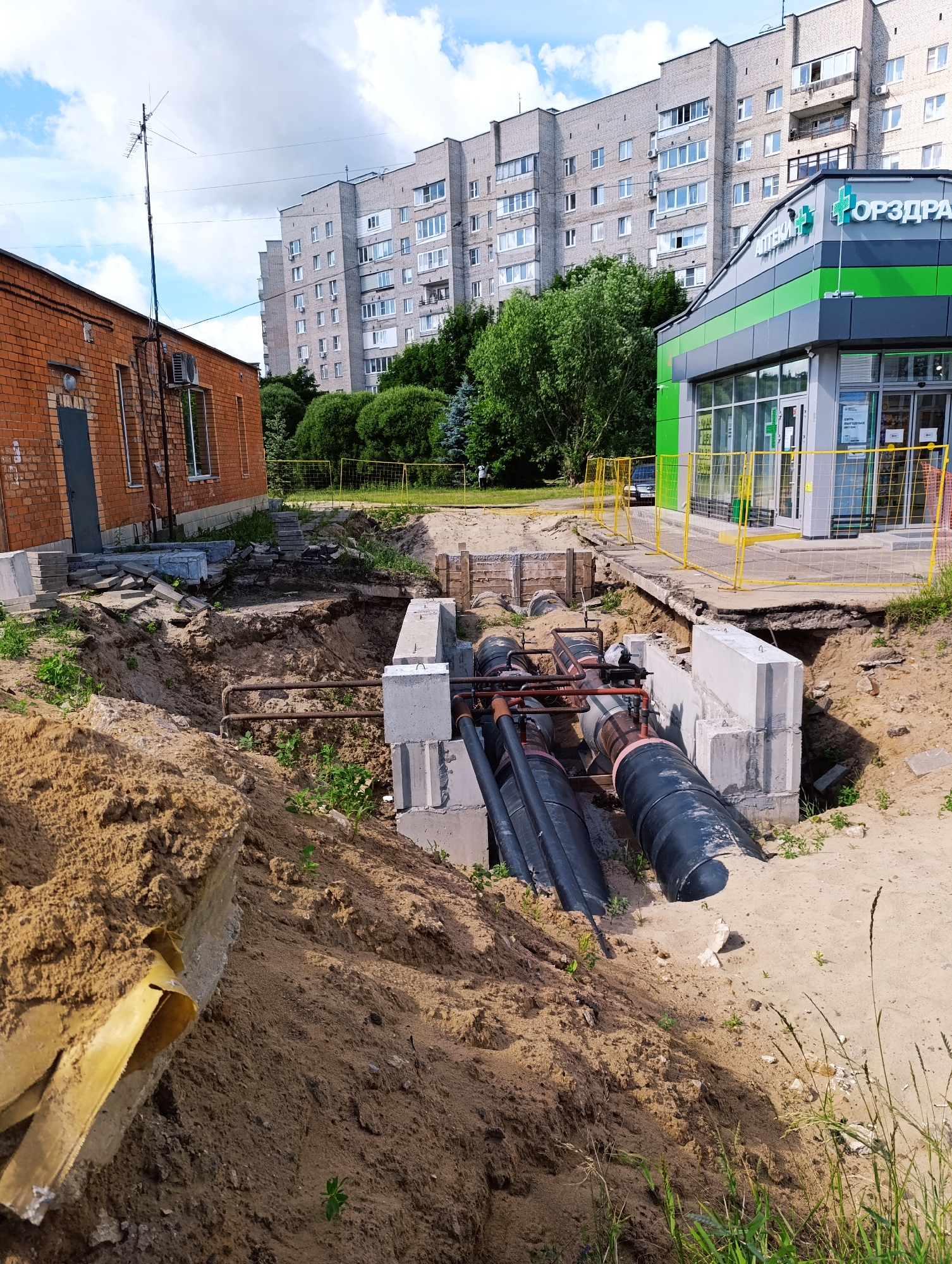 The image depicts an outdoor scene in an urban environment, possibly during daylight hours given the lighting. In the foreground, there is a large excavation pit filled with sand and soil, revealing exposed pipes. Several large, dark-colored pipes, likely for utilities, are visible within the pit, supported by concrete structures. Scattered construction materials, including concrete blocks and what appears to be insulation or protective sheeting, are present around the pit.
Behind the excavation, a brick building with a dark door and windows is visible to the left. Further in the background, a multi-story residential building with a grey facade and numerous windows dominates the skyline. To the right, a modern commercial building with a green and grey exterior, featuring large windows and signage, is partially visible. The signage on this building includes text in Cyrillic, with "АПТЕКА" (Apteka, meaning Pharmacy) and what appears to be a brand name "ОРЗДРА" (Orzdrav). A yellow safety fence encloses the area around the commercial building, separating it from the construction site. The sky above is partly cloudy, with patches of blue. Sparse vegetation, including grass and small plants, grows around the edges of the excavation and in the surrounding areas. No people are clearly visible in the immediate vicinity of the construction site, though a blurred figure can be seen in the far background near the commercial building. The scene suggests ongoing infrastructure work or construction in a residential and commercial area.