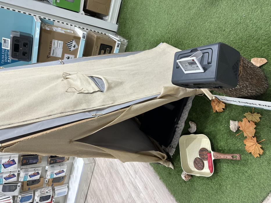 This image captures an indoor retail or exhibition display, rotated 90 degrees counter-clockwise, designed to simulate a camping scene.
The central subject is a beige, tent-like structure, possibly a sleeping bag or a prop, with an opening revealing a black television screen inside. Next to the tent, on artificial green turf, sits a dark grey portable power station. Attached to the power station is a small transparent holder with a label displaying information such as "299Wh" and "100W," along with the Greek text "φορητός σταθμός ενέργειας" (portable power station). The power station itself has visible ports and a textured casing.
Scattered on the artificial turf around the tent and power station are several dry autumn leaves and small rocks, enhancing the outdoor theme. In the foreground, a light-colored grill pan with a wooden handle contains two dark, round food items (resembling burgers or sausages) and a red chili or pepper.
In the background, bright white retail shelving units are visible. On these shelves, several cardboard boxes are stacked, prominently featuring logos for "BLUETTI" and "ECOFLOW," indicating they are likely portable power generators. Some boxes also display hazard symbols. Further to the left, more shelving units are packed with various consumer electronics, including phone accessories, with text like "GLASS" visible on some packaging. A large white sign in the background partially reads "ΓΙΑ ΝΑ ΕΧΕΙΣ ΕΝΕΡΓΕΙΑ ΠΑΝΤΟΥ" (To have energy everywhere). The flooring transitions from artificial turf to light-colored tiled floor at the bottom of the image.