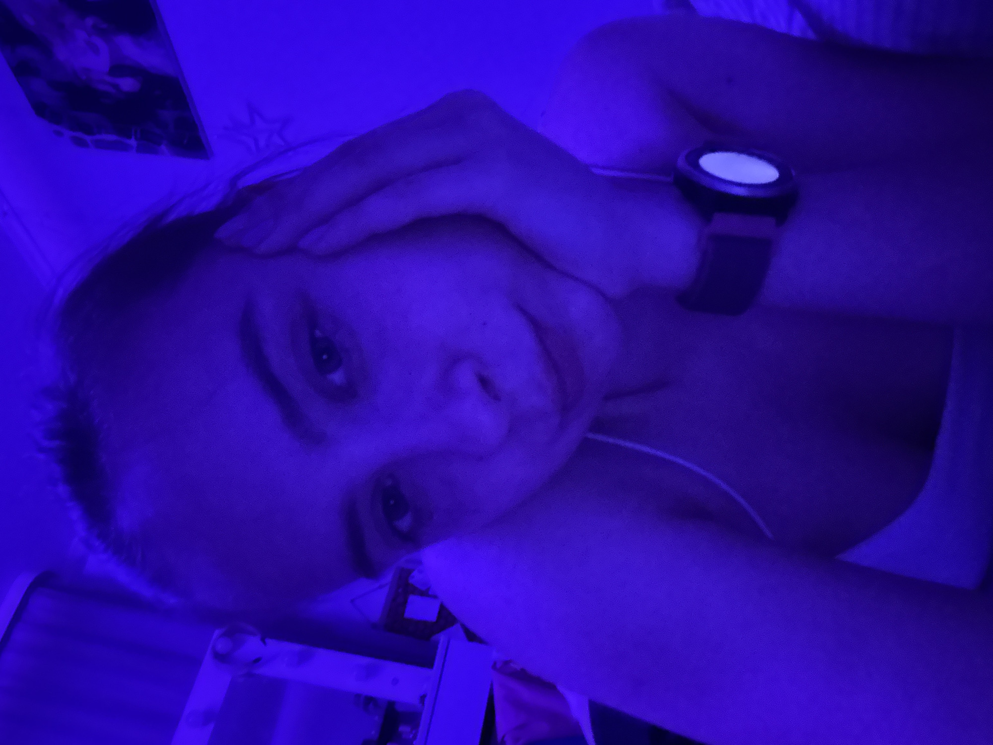 A woman lies on a bed in a dimly lit room, possibly in Прикамский, Russia. The predominant color is blue. She is wearing a watch and resting her head on her hand. A white tank top is visible. In the background, there is a shelf or table and a picture on the wall.