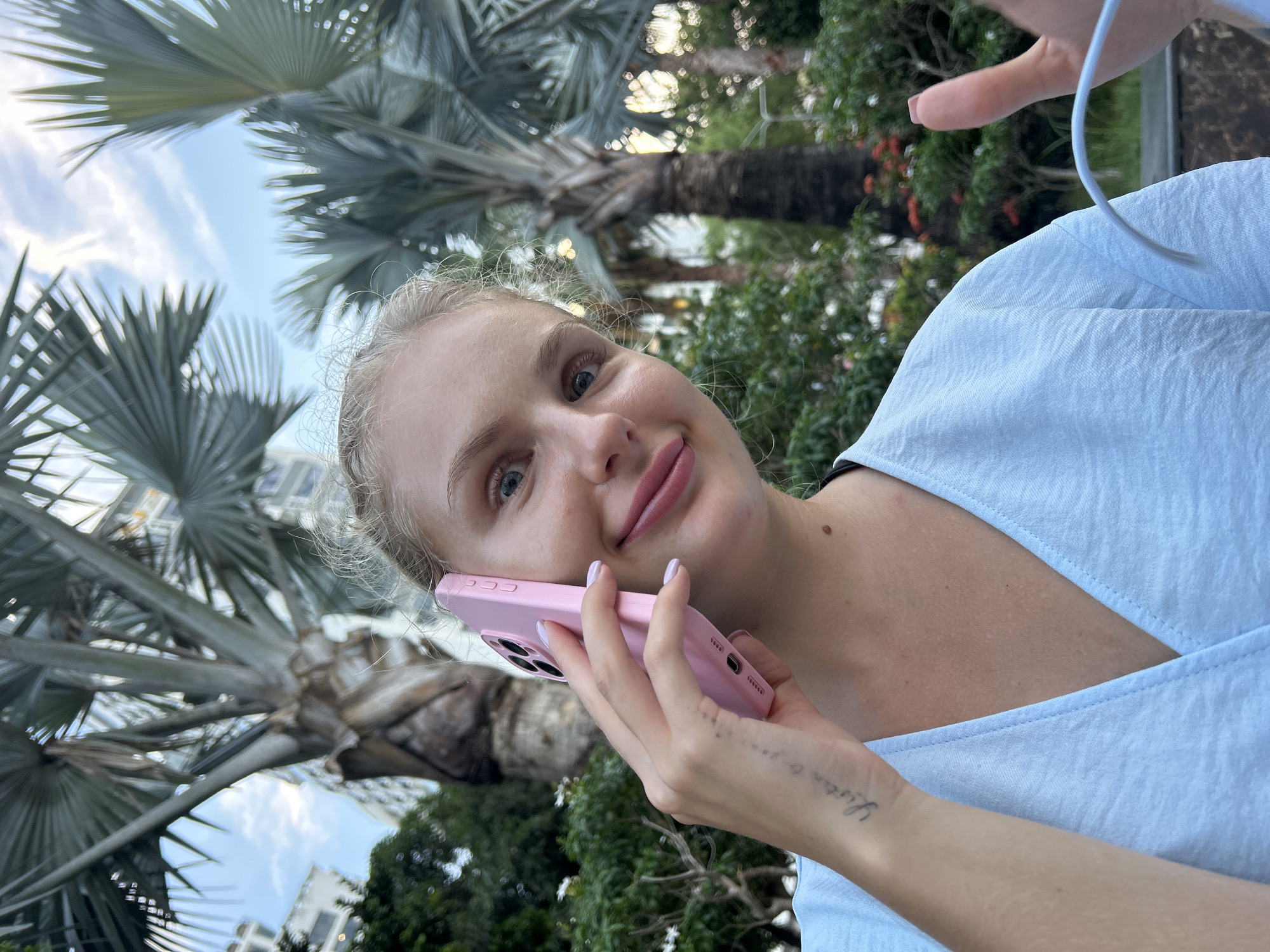 An outdoor, medium shot shows a young woman holding a pink phone to her ear. She has blonde hair tied up, blue eyes, and rosy lips. She is wearing a light blue, V-neck top. Her left arm is extended, and a tattoo is visible on her inner wrist. She is in a park-like setting with palm trees and lush green foliage. The background features buildings and a blue sky with scattered clouds. The lighting suggests it is late afternoon or early evening. The location is specified as Nha Trang Ward, Vietnam.