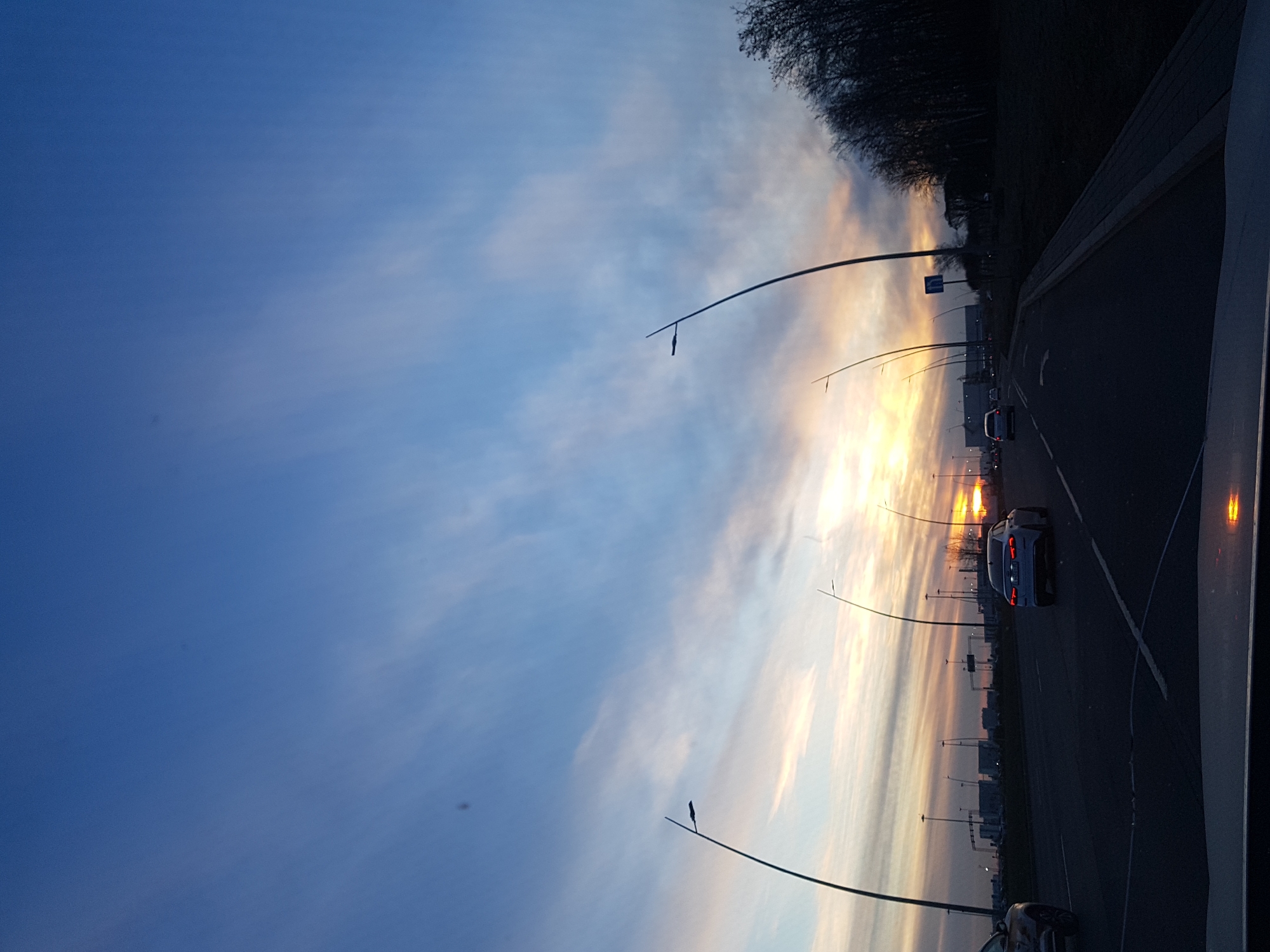 The image shows a view from inside a car driving on a road, likely in Saint Petersburg, Russia, as suggested by the architectural style visible in the background. It is dusk or dawn with the sun low on the horizon, creating a vibrant orange glow behind a bank of clouds. Streetlights line the road, and several cars are visible. A dark tree line is present on the right side of the road. The sky is predominantly blue, fading into the colors of the sunset near the horizon.