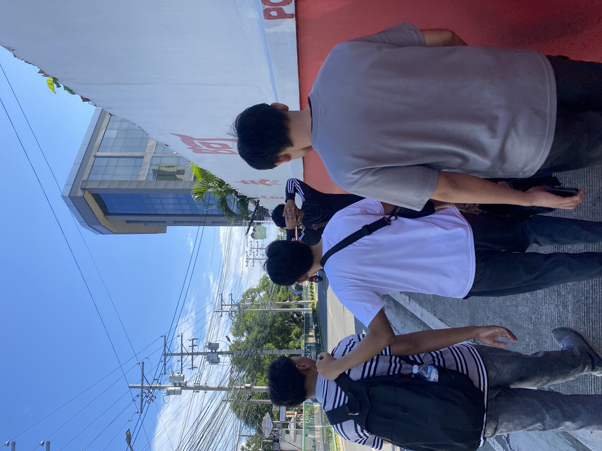In Davao City, Philippines, four young men walk along a sidewalk next to a red and white wall. The man in the foreground is wearing a grey t-shirt and dark pants. To his left, a man in a white t-shirt and black pants walks with a black bag slung over his shoulder. To the left of them, a man in a striped shirt and grey pants carries a black backpack. The fourth man, partially obscured, is wearing a black hoodie and has his hands over his eyes. Behind them, utility poles with a dense network of wires stretch across the bright blue sky. A modern, glass-fronted building is visible in the distance, as are trees and a road with a single motorcycle. The wall has red characters and a partial "PO" visible.