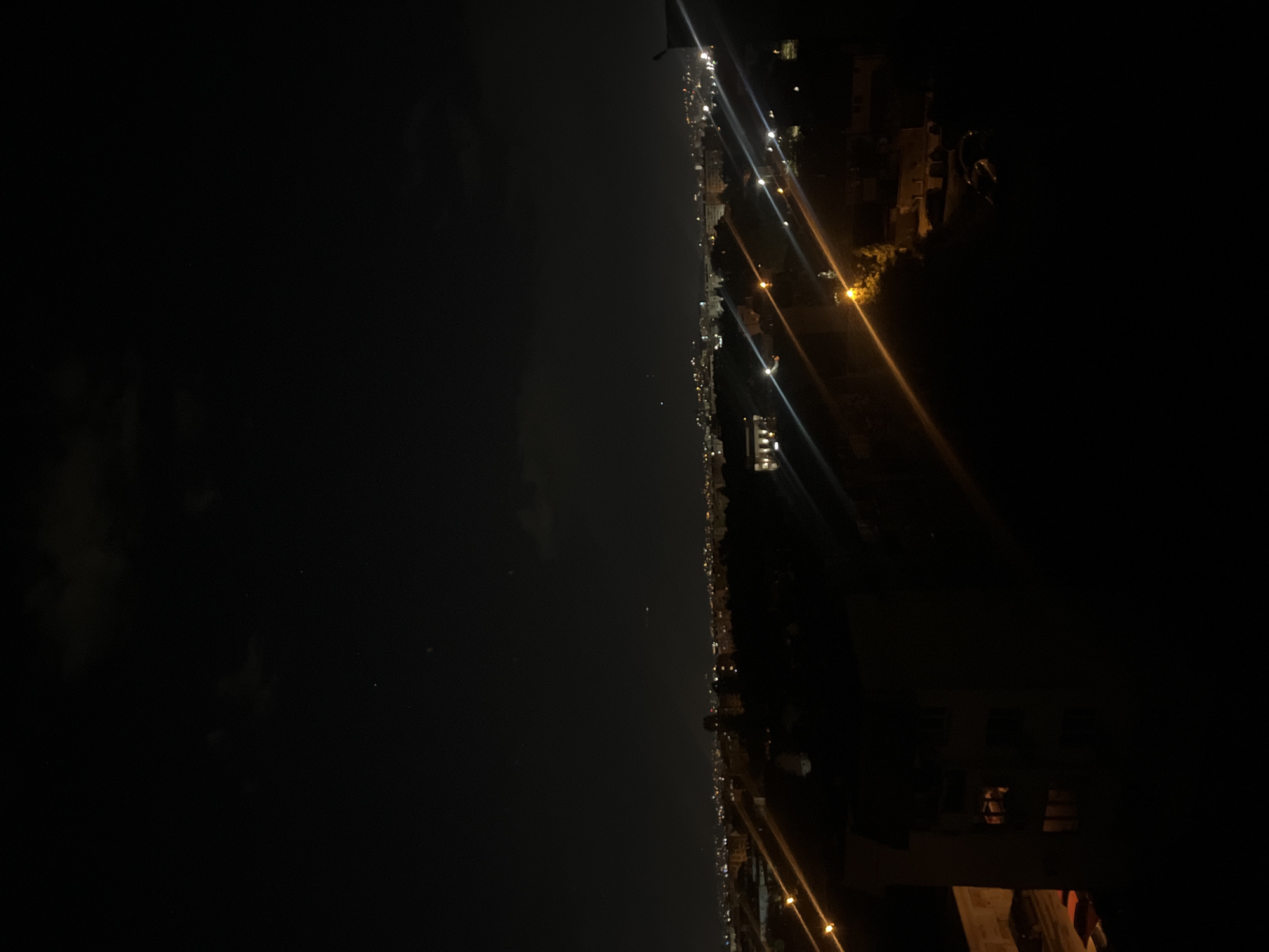The image shows a nighttime view of a city skyline, characterized by a dark sky and bright lights stretching across the horizon. Street lights and building lights create a dense, illuminated line in the middle of the frame, indicating an urban area. The lower portion of the image shows a dark foreground, suggesting the viewer is positioned in a building or elevated location overlooking the city. The exact location in Sofia, Bulgaria cannot be determined from the image. No people, actions, or interactions are directly visible in the image.