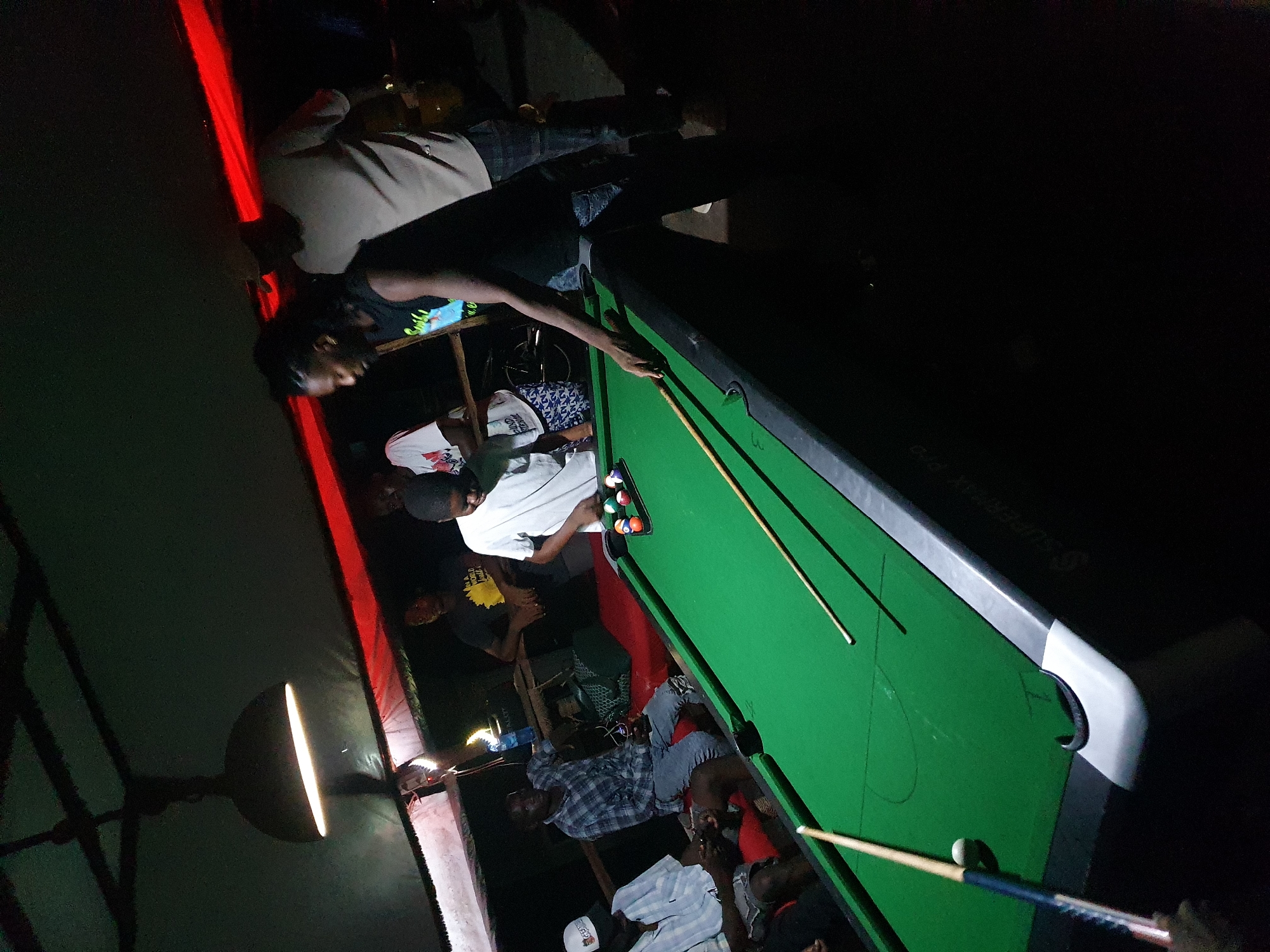 People are playing a game of pool on a green pool table indoors. The location appears to be dimly lit, with a red light source in the background. Around the table, individuals are standing and observing the game, while some are seated. Pool cues and balls are visible on the table. The pool table has a silver trim. The place is likely a bar or a recreational facility in Lokoja, Nigeria.
