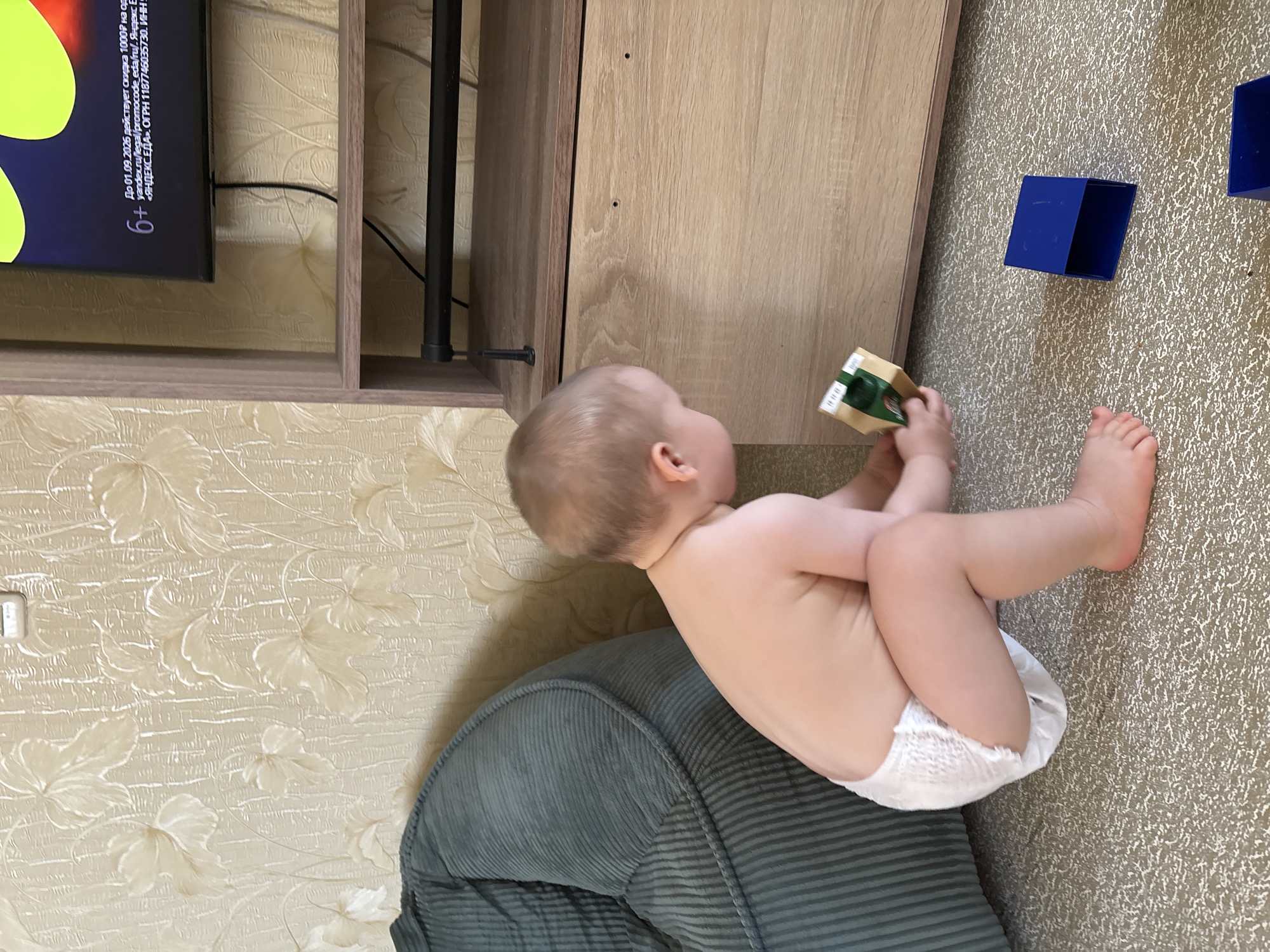 In this medium shot, a young baby, likely a toddler, sits on a textured, grey upholstered couch. The baby is wearing only a diaper and is facing away from the camera, looking towards a wooden cabinet. The baby is reaching out and holding a small, rectangular object with green and brown colors. The cabinet is light brown with a subtle wood grain pattern and has visible hardware and screw holes. To the right of the cabinet, on a speckled beige carpet, are two bright blue, cubical objects. In the background, the wall is covered in a cream-colored wallpaper with a raised, leaf-like pattern. To the left of the scene, a portion of a black television screen is visible, displaying a yellow graphic and some text. The text on the screen is in Russian and indicates a promotional offer. The lighting suggests it is daytime, and the indoor setting appears to be a living room or play area. The baby's posture and focus on the object suggest an activity of exploration and play.