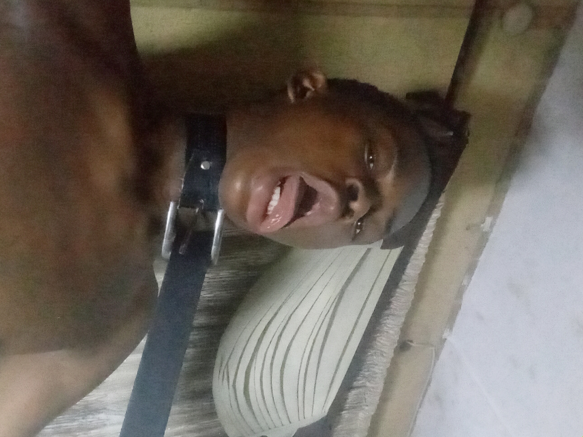 A person with dark skin is lying on what appears to be a bed or pallet, with their head tilted to the side and their mouth open, tongue protruding. A black leather-like collar with a metal buckle is around their neck. The background appears to be a simple room with bare or unfinished walls, possibly in Nkpor, Nigeria, based on the prompt.