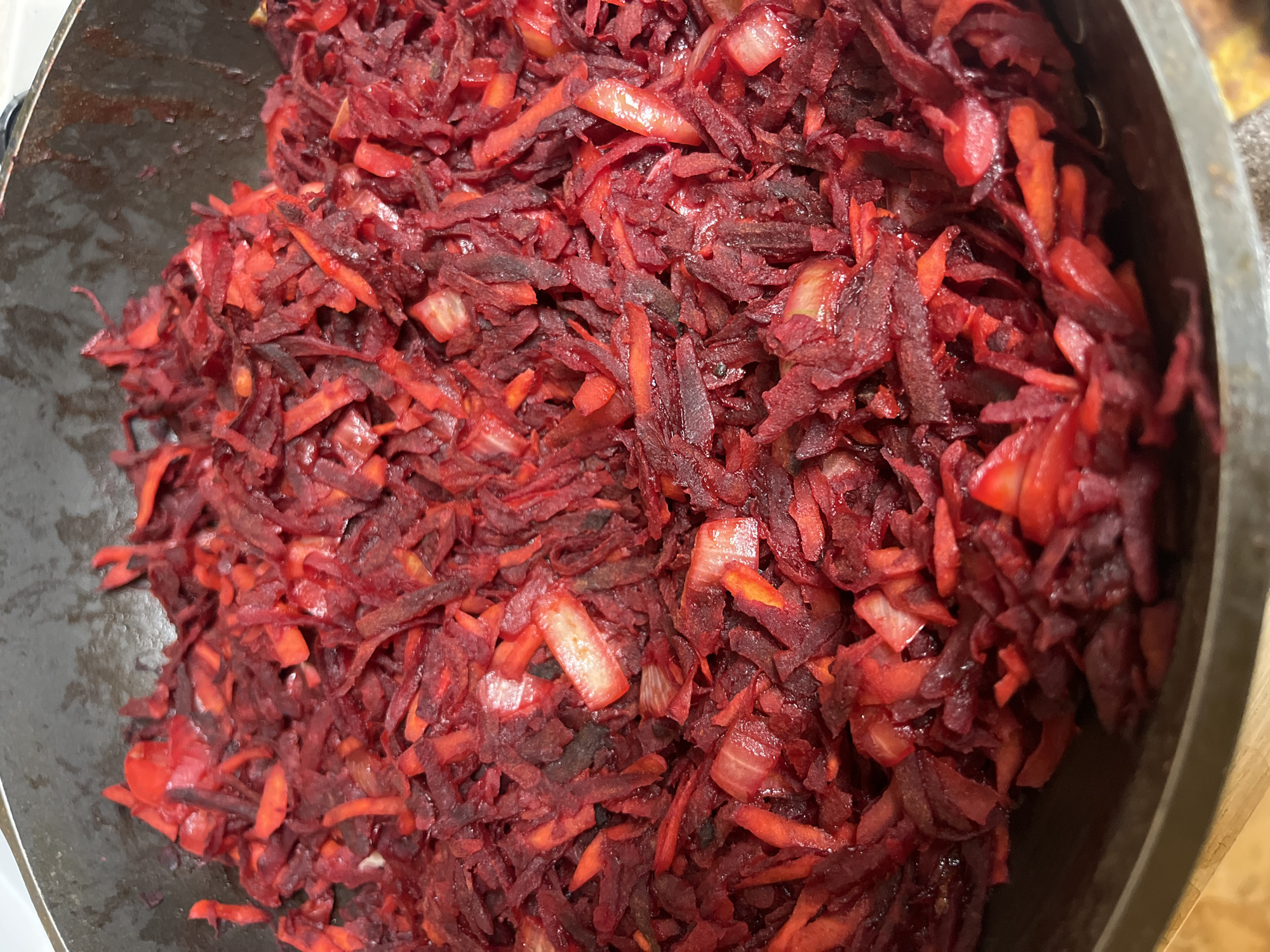 A close-up, overhead shot shows a dark, textured metal pan filled with shredded beets and some lighter-colored ingredients, possibly onions. The beets are a deep, vibrant red, with some pieces appearing darker and more cooked than others. The shredded vegetables are piled high in the pan, filling most of the frame. The lighting is bright, casting some subtle shadows and highlighting the glossy texture of the cooked vegetables. The pan itself appears to be a stovetop pan, with a slightly worn, dark surface. The background is out of focus, but appears to be a wooden surface. The scene is likely a kitchen or cooking environment. No people are visible in the image. The specific location in the city of Vasilyevo, Russia, cannot be determined from the image.