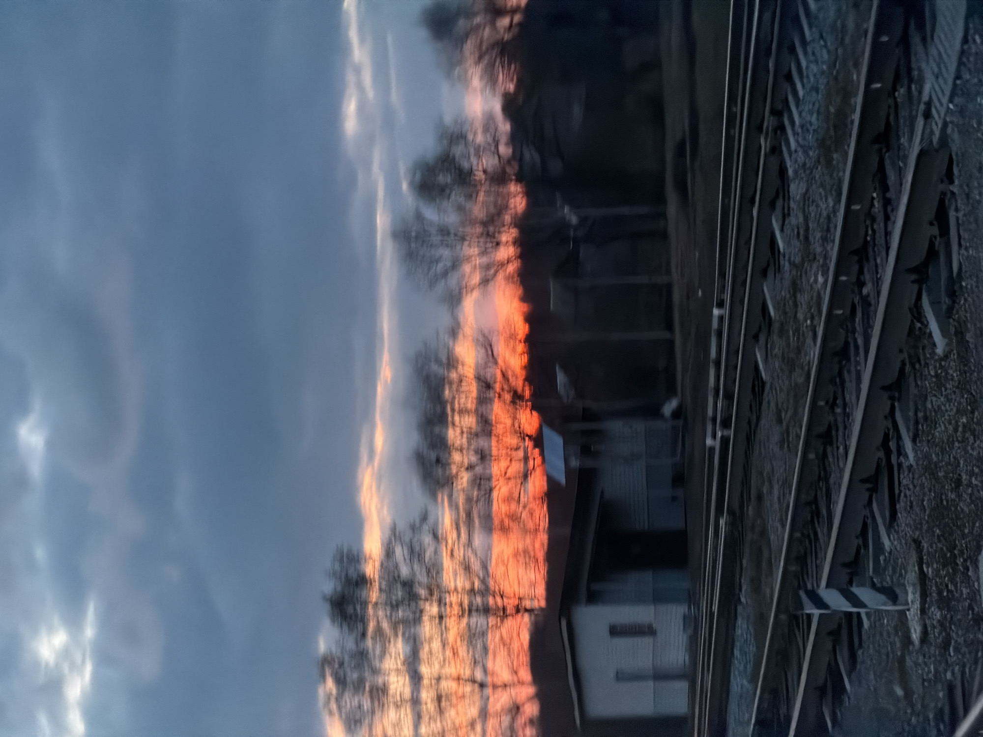 The image, appearing rotated vertically but described as if oriented horizontally, depicts a serene sunset or sunrise scene in Кварса, Russia. The sky dominates the upper part of the frame, showcasing a beautiful transition from vivid orange, red, and yellow hues near the horizon to softer blues and greys higher up, with scattered clouds catching the light.
Silhouetted against the colorful sky are numerous bare, deciduous trees in the mid-ground, their intricate branch patterns prominent. Below the horizon, several low-rise buildings, likely residential houses, are visible, partially obscured by the trees and shadows. One building, closer to the center-left, features a light-colored facade and a dark roof.
To the right, a set of railway tracks runs diagonally from the foreground into the distance, laid on a bed of gravel ballast. A small, striped black and white marker stands upright next to the tracks in the lower-right foreground. The low angle of the sun and the resulting shadows suggest either dawn or dusk. No people are visible, and there is no discernible text in the image. The bare trees indicate a season of late autumn, winter, or early spring. The scene conveys a quiet and atmospheric moment.