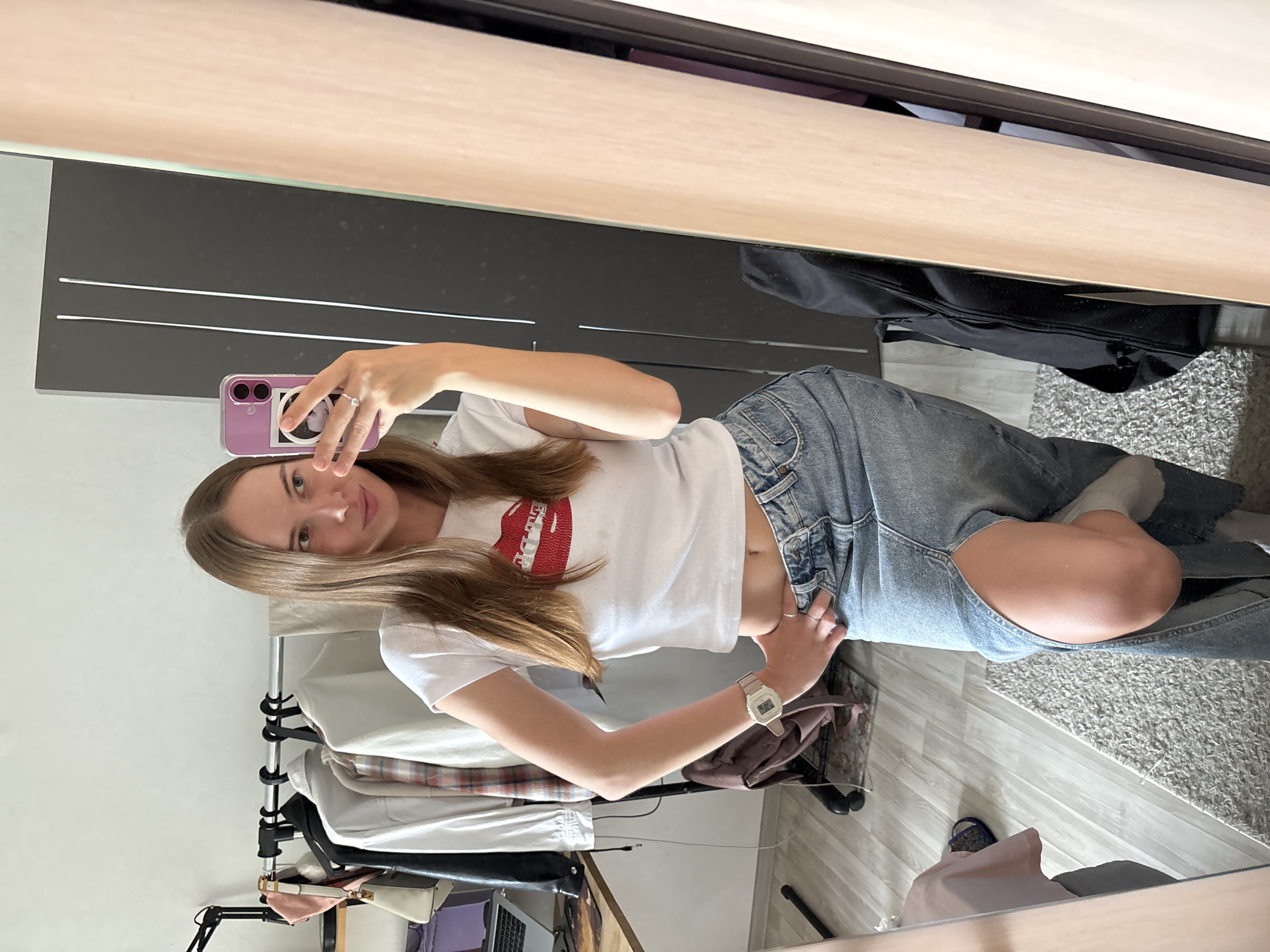 In a domestic indoor setting, a young Caucasian woman with long brown hair, wearing a white crop top with a red sequined lip design and a denim maxi skirt with a high slit, is taking a mirror selfie. She is standing in what appears to be a bedroom or dressing area, with a clothing rack full of clothes visible behind her to the left, a desk with a laptop, and a full-length mirror to her right. A black duffel bag is visible on the floor near the mirror. The floor is covered with light-colored laminate flooring and a textured gray rug. The overall impression is of a casual, personal moment captured in a home environment. The image does not contain any specific visual cues that can be used to identify the location as the city of Perm, Russia.